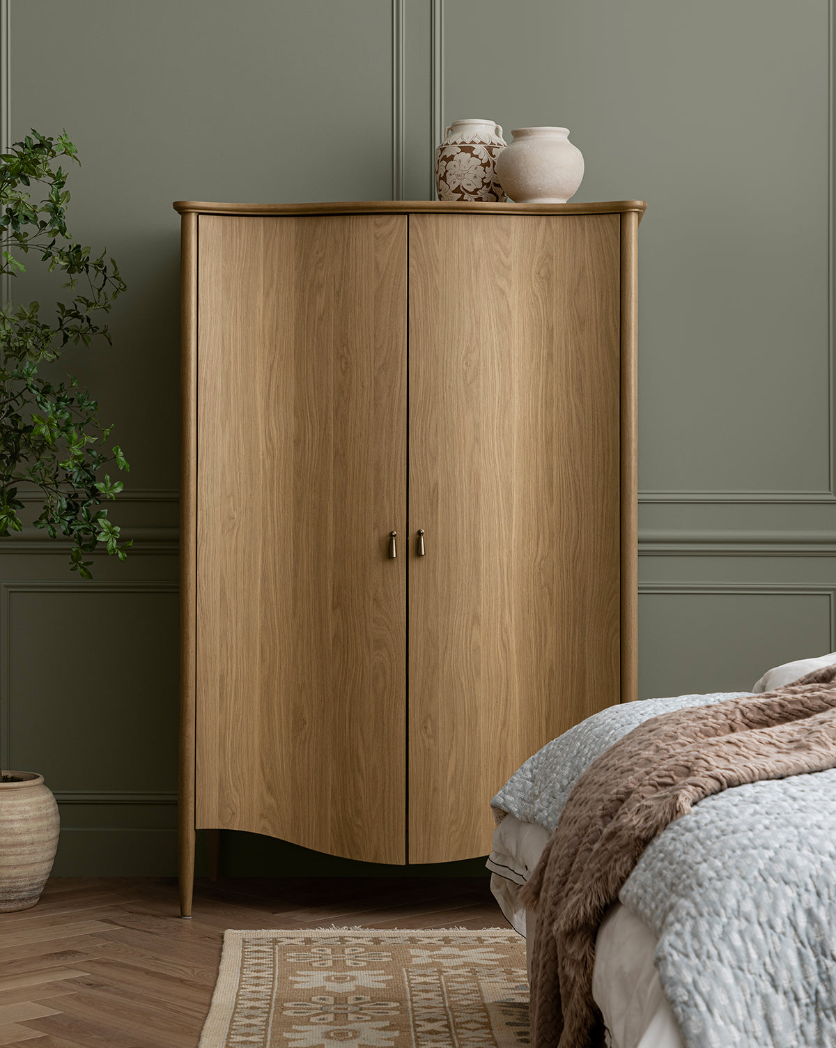 A light wood cabinet with two doors stands against a sage green paneled wall. On top sits the Nola Paper Mache Vase and another decorative piece. A plant, bed with textured blankets, and a patterned rug are partially visible in the room, by McGee & Co.
