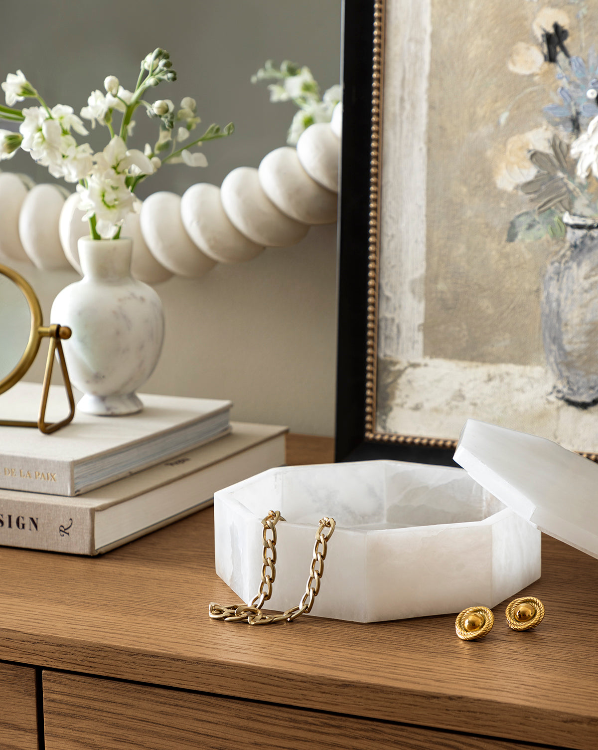 The Sonya Alabaster Box, octagonal in shape, sits open on a wooden dresser next to stacked books, a vase with white flowers, and a framed painting. Gold chain and earrings rest inside the elegant box.