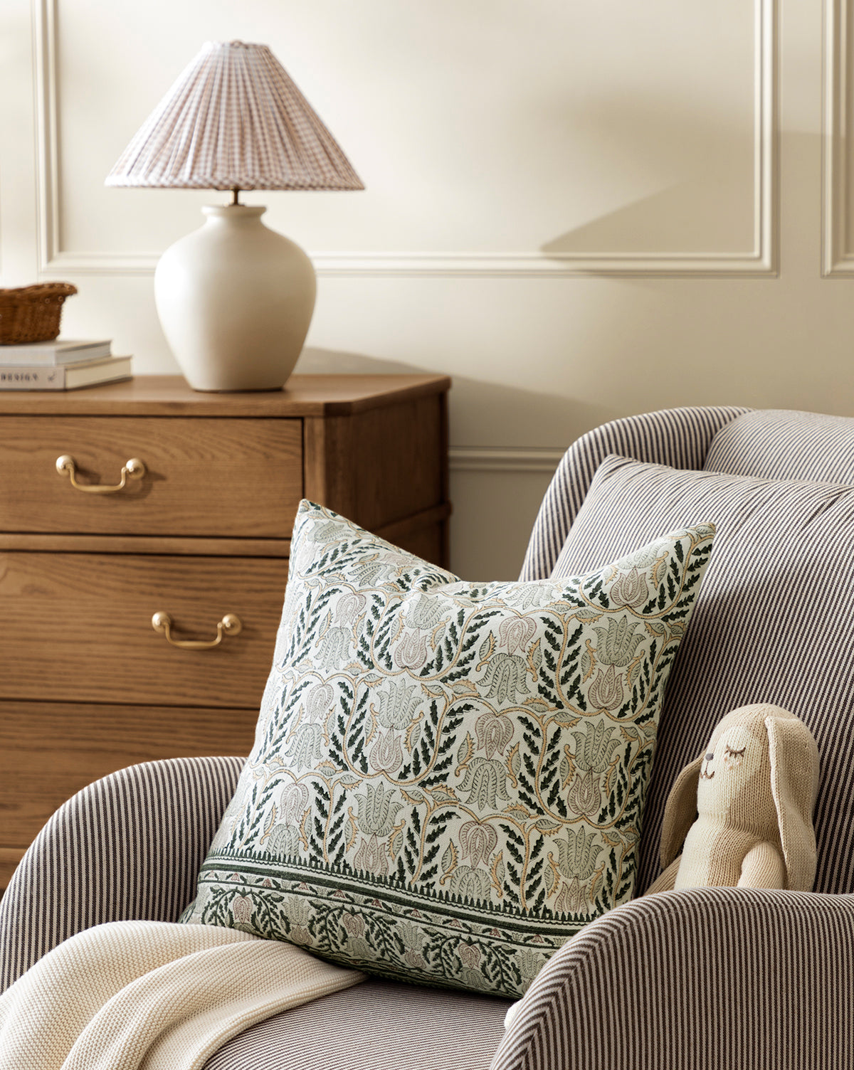 An armchair with the Ellsbeth Pillow Cover sits beside a wooden dresser and a lamp with a pleated shade in a cozy, softly lit room, by McGee & Co.