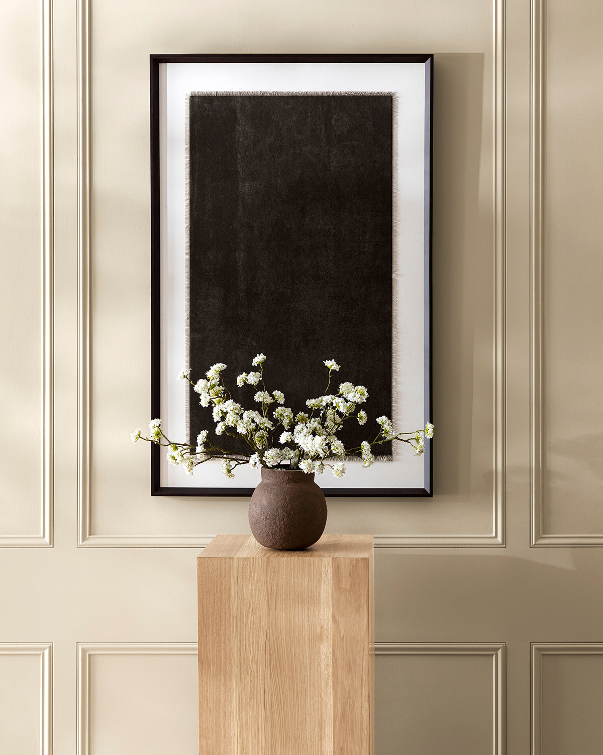 A minimalist interior showcases the Frederick Paper Mache Vase with white blossoming branches on a wooden pedestal, set before a beige paneled wall adorned with framed abstract black art, by McGee & Co.