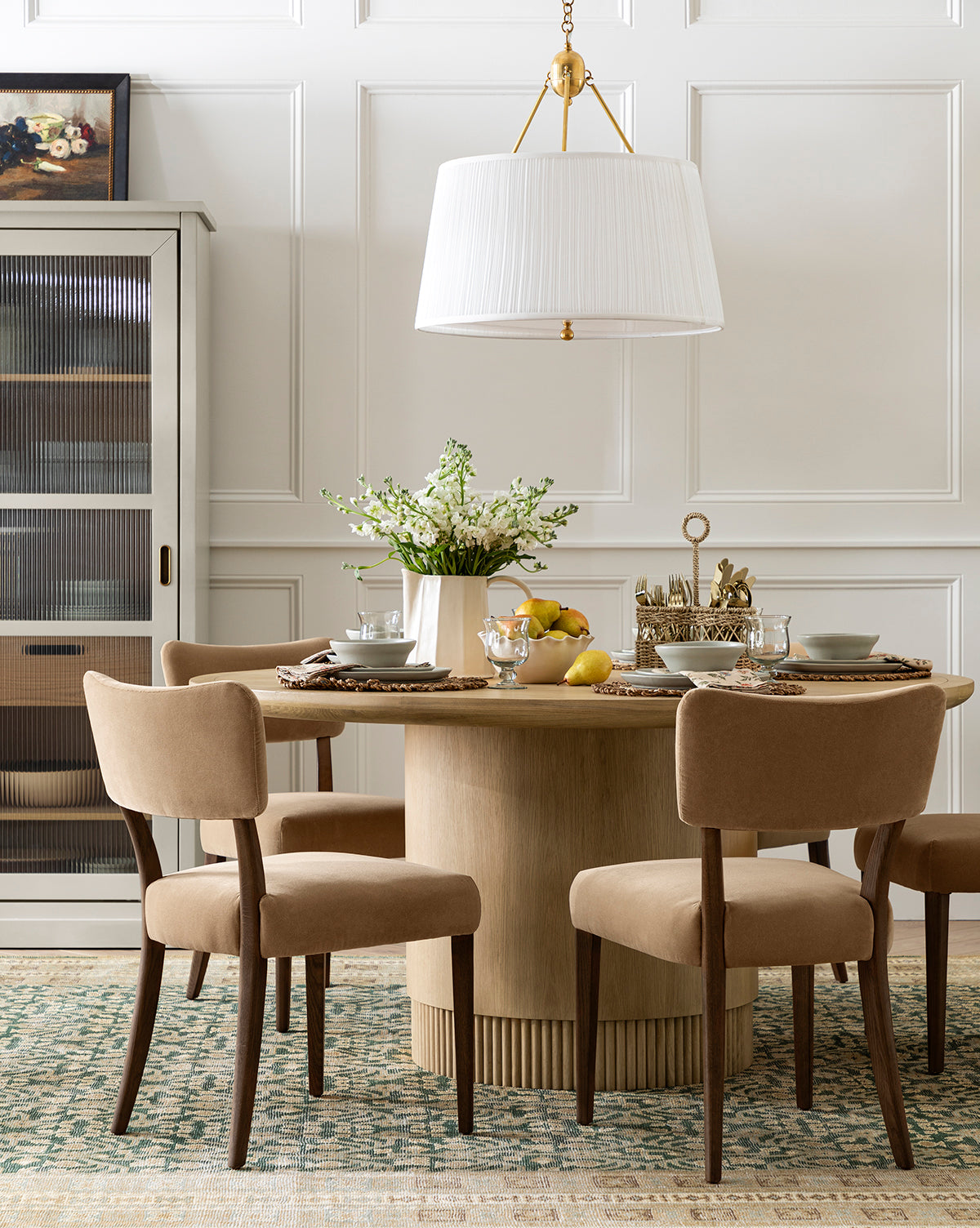 A dining room with a Gilroy Round Oak Dining Table with a reeded base, four beige upholstered chairs, a large white pendant light, elegant tableware, a patterned rug, and cabinet create this neutral-toned heritage oak finish setting by McGee & Co.