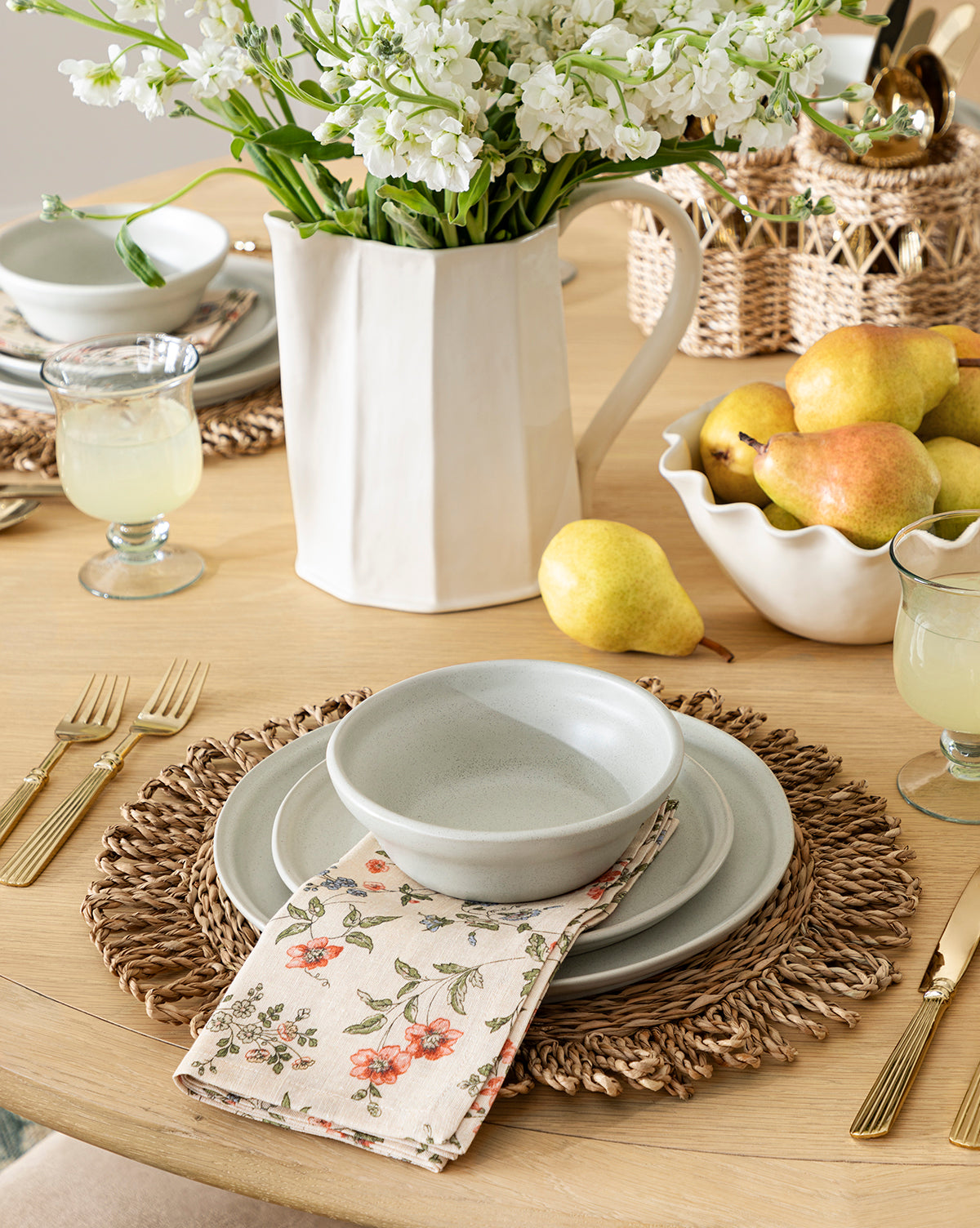 A wooden table set for a meal with woven placemats, Aiden Soft Gray Dinner Plates and bowls, floral napkins, gold cutlery, glasses of lemonade, a vase of white flowers, and a bowl of pears, by McGee & Co.
