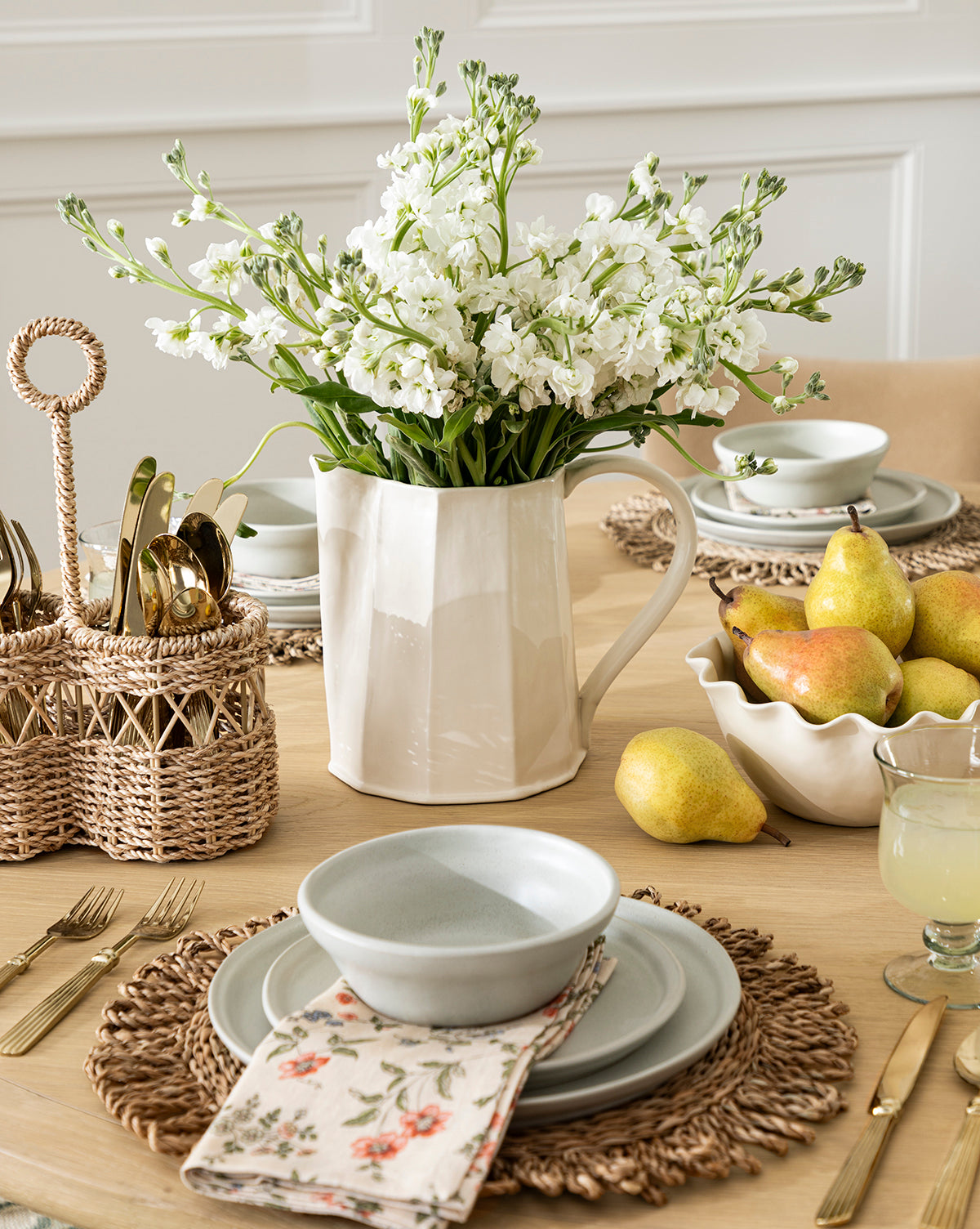 A dining table set for a meal features gray dishes, gold cutlery in a basket, a floral napkin, rattan placemats, the Fielding Ceramic Pitcher with white flowers, fresh pears, and lemonade for cozy rustic kitchen decor, by McGee & Co.