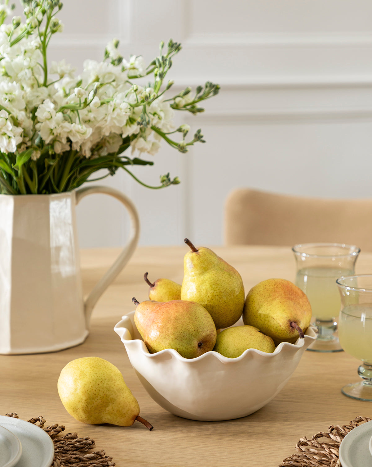 An Olindo Serving Bowl from the Olindo Serving Bowls (Set of 2) in cream, filled with pears, rests on a wooden table beside two juice glasses and a white ceramic pitcher with white flowers by McGee & Co.