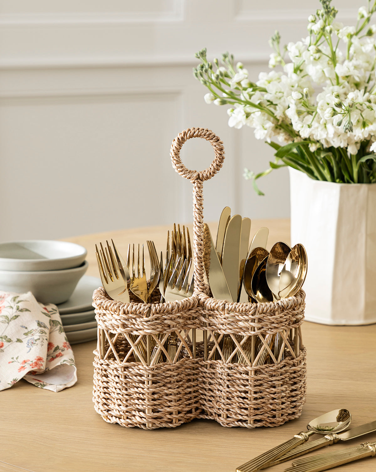 The Stokes Woven Caddy organizes gold-toned forks, knives, and spoons on a wooden table with stacked dishes, a floral napkin, and a white vase of flowers—ideal for adding style to any kitchen counter, by McGee & Co.