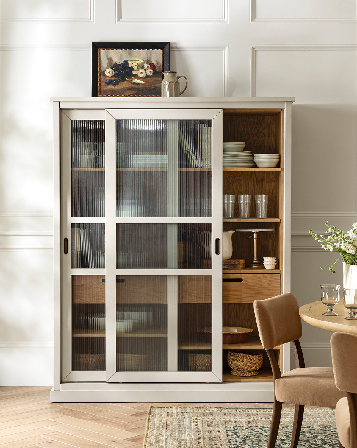 A white cabinet with ribbed glass doors showcases kitchenware, while Tableside by Joyce Washor, plus a jug and cup, adorn the top. A set dining table and chairs are arranged in the foreground, by McGee & Co.