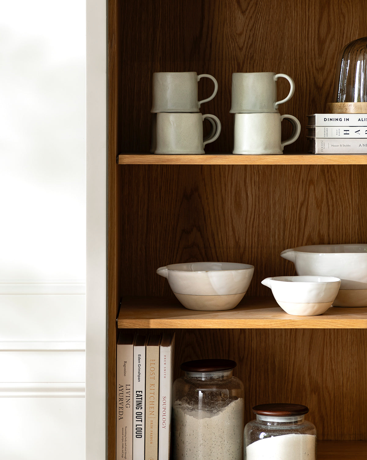 A wooden kitchen shelf displays stacked mugs, white ceramic bowls, vintage baking books, two glass jars with dry ingredients, a set of Karina Stoneware Batter Bowls (Set of 4), and a stack of plates against a warm wood grain background, by McGee & Co.