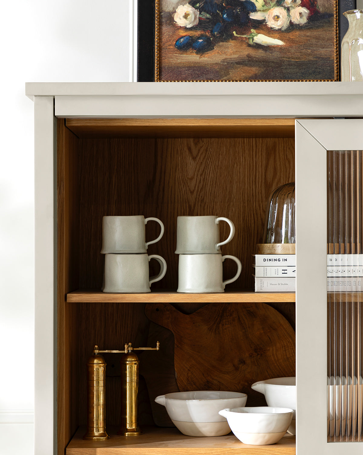 A cabinet with two shelves displays stacked white mugs, an Aiden Soft Gray Mug, books, a cloche, white bowls, a wooden board, and two brass salt and pepper grinders. A floral painting rests on top of the cabinet, by McGee & Co.