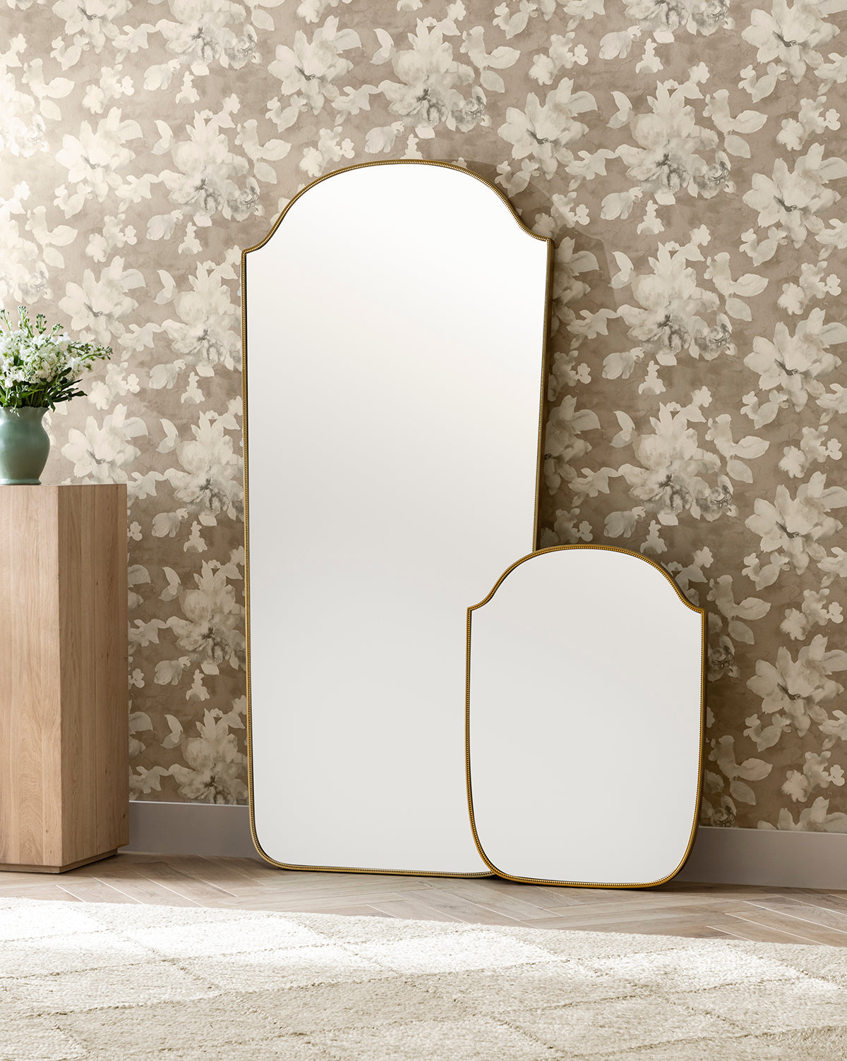 Two decorative mirrors, including the Sussex Floor Mirror with a beaded frame, lean against a floral wallpapered wall. On the left stands a wooden cabinet topped with a green vase and plant, while a beige rug covers the floor, by McGee & Co.