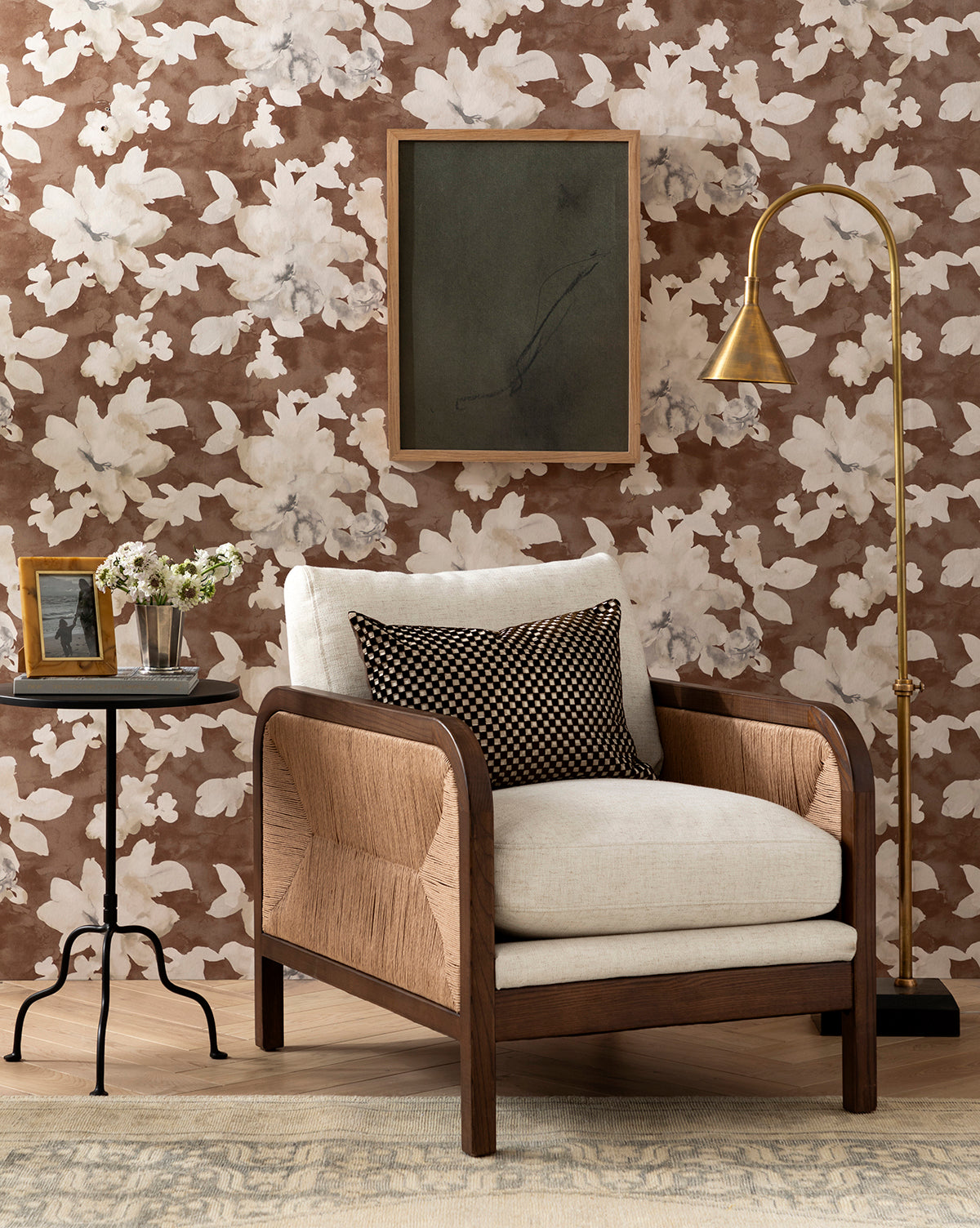 A Garvey Lounge Chair with plush cushions and a dark patterned pillow stands by a small round side table topped with framed photos and flowers, set against floral brown wallpaper beneath a gold floor lamp and an abstract painting by McGee & Co.