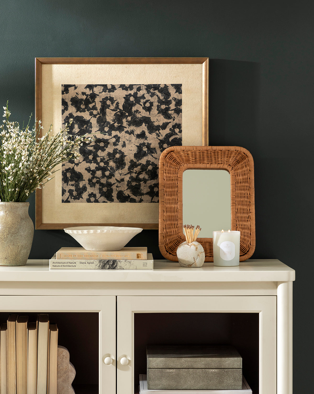 A cream cabinet with books, a ceramic vase of white flowers, stacked books, a round container with matches, a white candle, a rattan-framed mirror, and the Woodblocked Botanical art piece in a gold frame against a dark green wall.