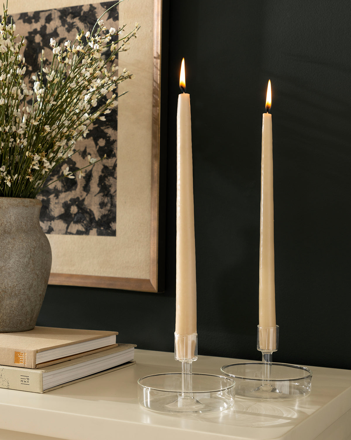 Two lit cream taper candles in clear Sofia Candle Holders sit on a white surface near stacked books and a vase of white flowers, with framed wall art in the background.