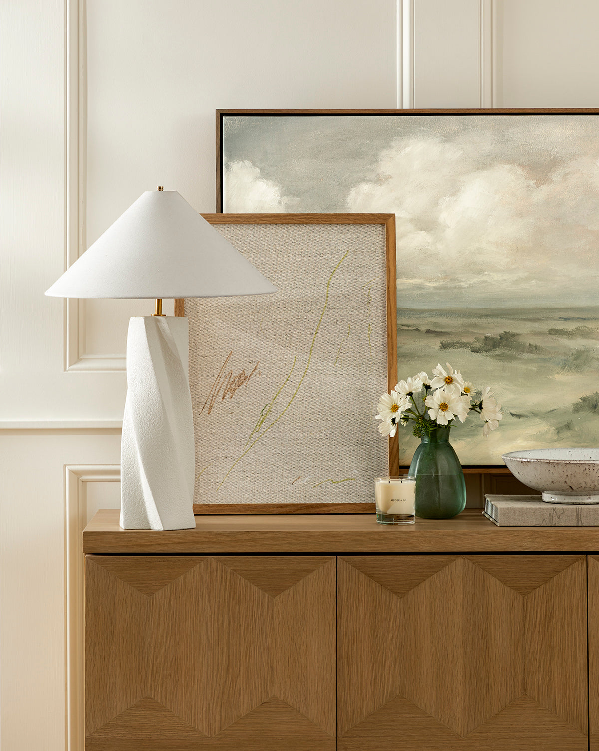 A wooden sideboard displays the modern Bettencourt Table Lamp with a linen shade, two framed art pieces, a small vase of white flowers, a candle, and a decorative bowl against a cream-colored paneled wall, by McGee & Co.