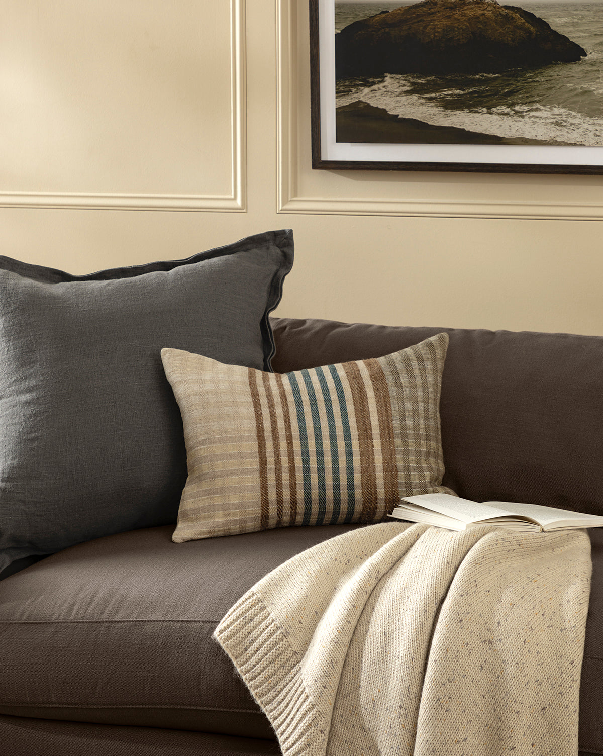 A brown sofa with a gray pillow, a striped beige pillow, the Makerspalm Otis Knit Throw in cream, and an open book. Beige walls and a framed rocky shore print hang above the sofa, blending seamlessly with pieces from McGee & Co.