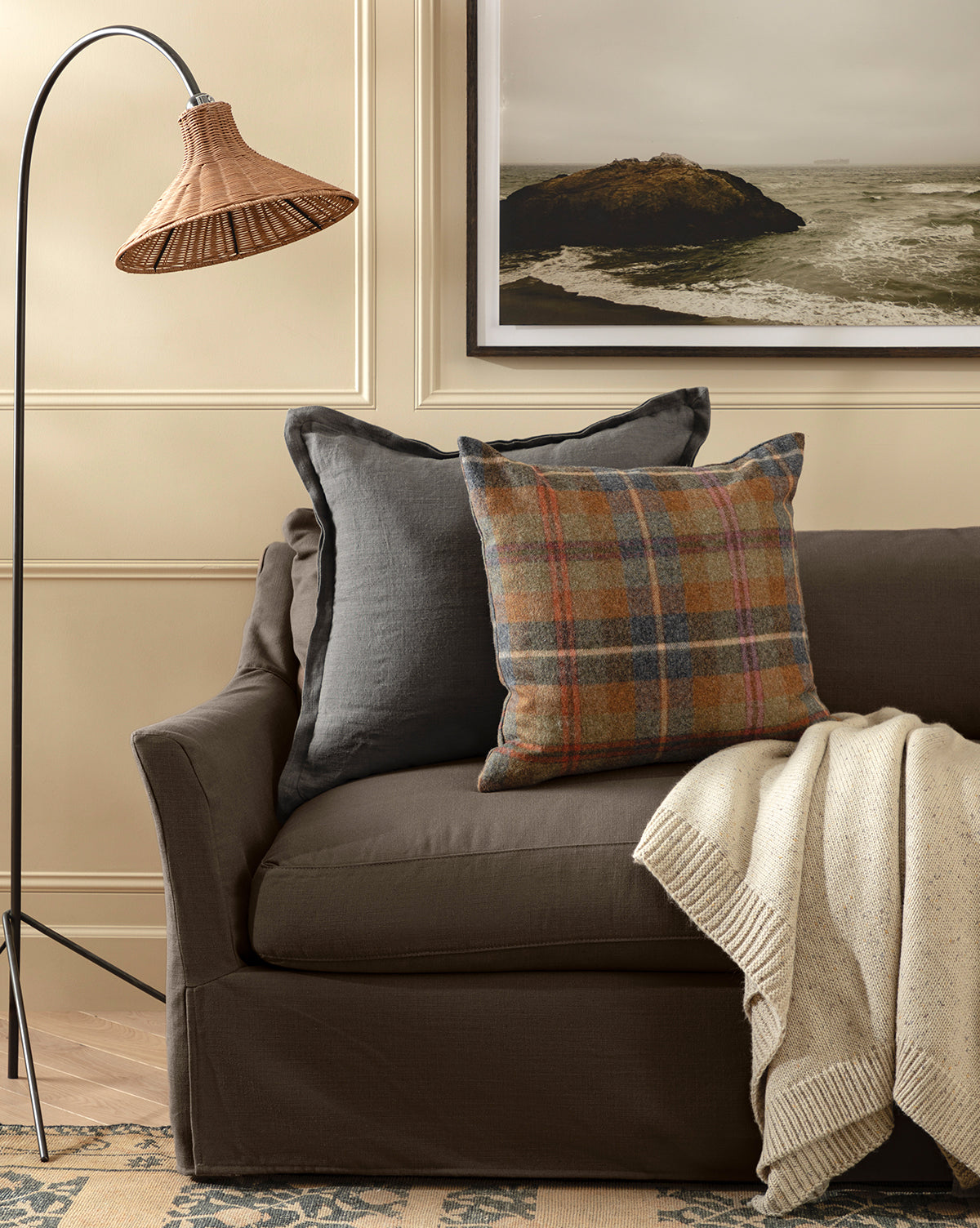 A cozy living room scene with a brown sofa, Liam Double Flange Pillow Cover, plaid pillow, cream knit throw, woven floor lamp, and framed coastal landscape on the wall, by McGee & Co.