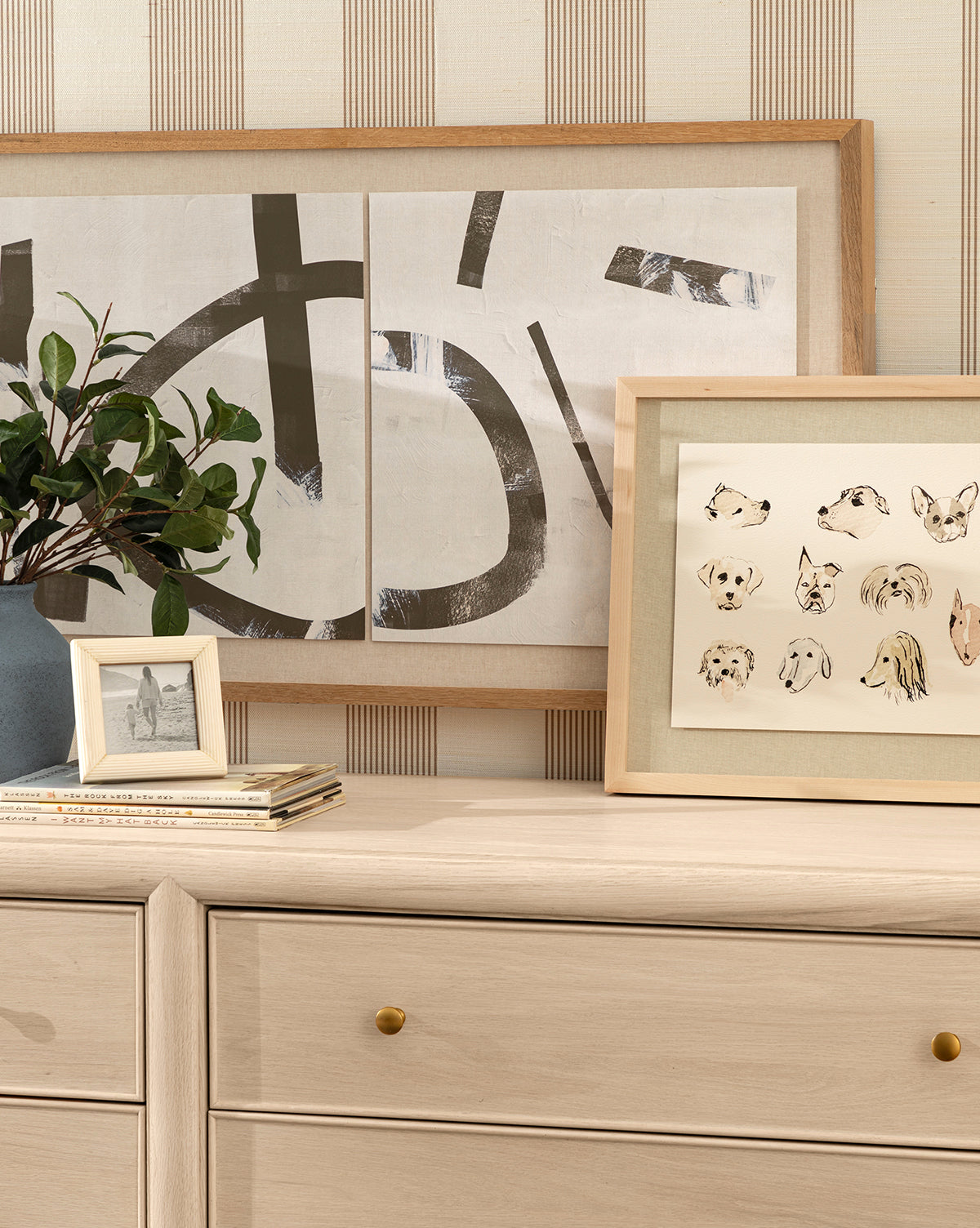 A light wood dresser with gold knobs displays Woof framed wall art, a dog sketch, a small photo in a blue frame, stacked books, and a green leafy plant against striped wallpaper by McGee & Co.