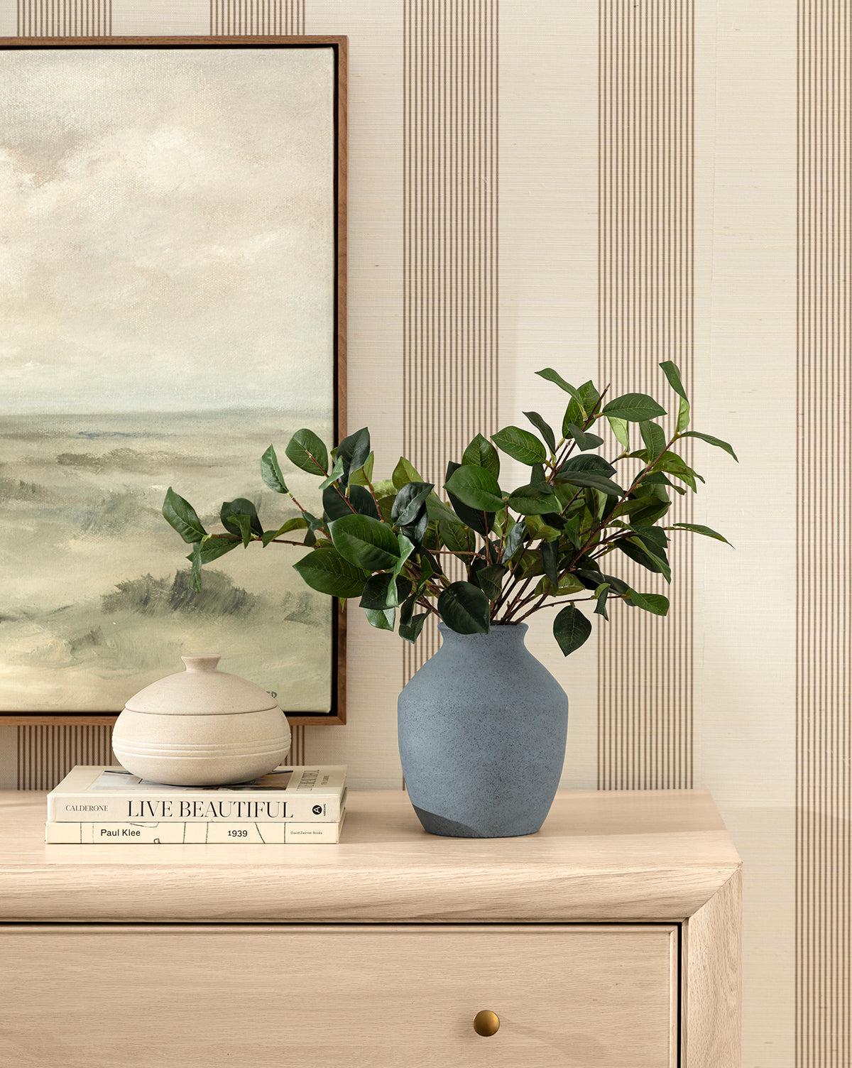 The Delgado Vase, filled with green leafy branches, rests on a light wooden dresser beside a small white ceramic jar. Behind them are stacked books and a landscape painting, all set against striped wallpaper, by McGee & Co.