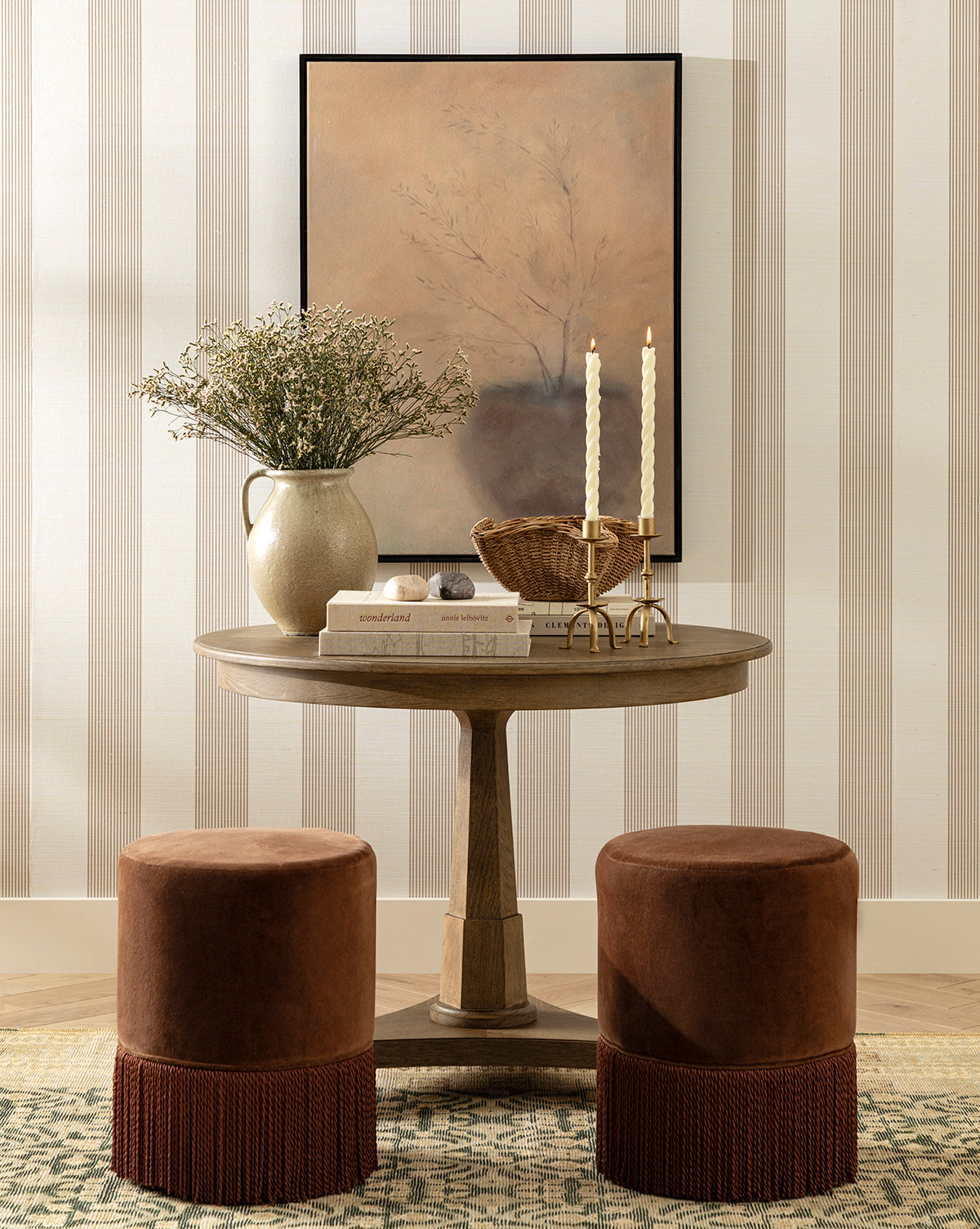 The Marna Entry Table is paired with two brown velvet stools, a vase of greenery, books, a basket, and two tall candles. Neutral striped wallpaper and framed abstract art complete the stylish look by McGee & Co.