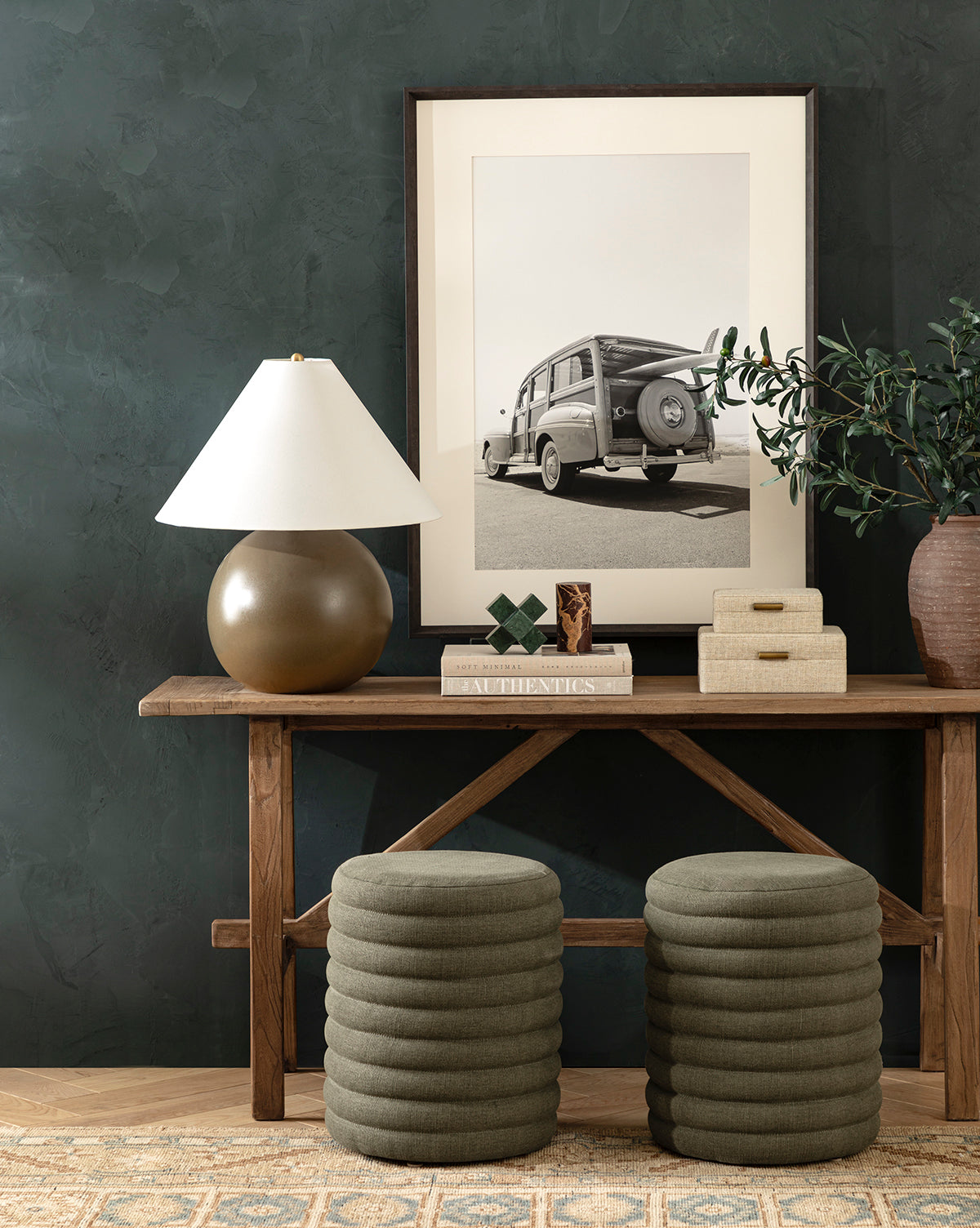 A wooden console table topped with a bronze lamp, books, decor, and a potted plant stands against a dark green textured wall beneath a framed black-and-white vintage car photo. Two Made Goods Jefferson Stools in green quilted fabric sit underneath. McGee & Co.
