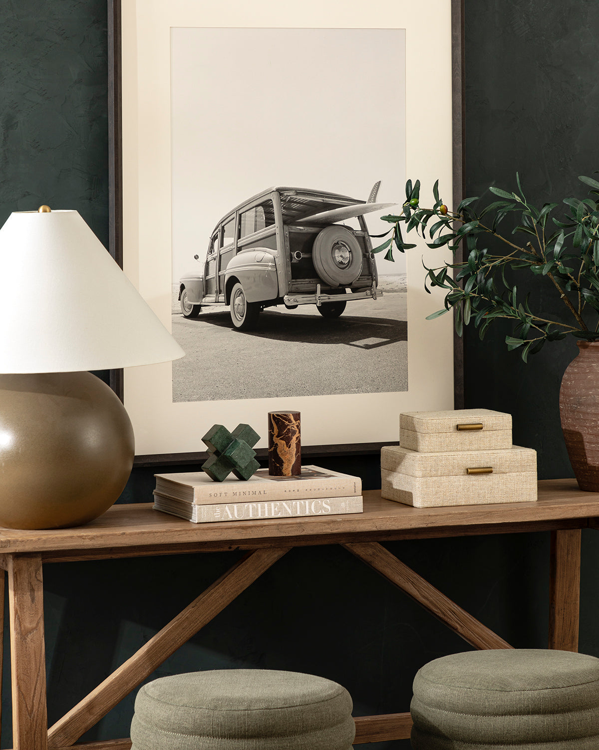 A wooden console table with books, the Diani Marble Cuboid Object, and a lamp sits beneath a large black-and-white vintage car photo. Green stools and a potted plant complete this stylish, cozy setting.