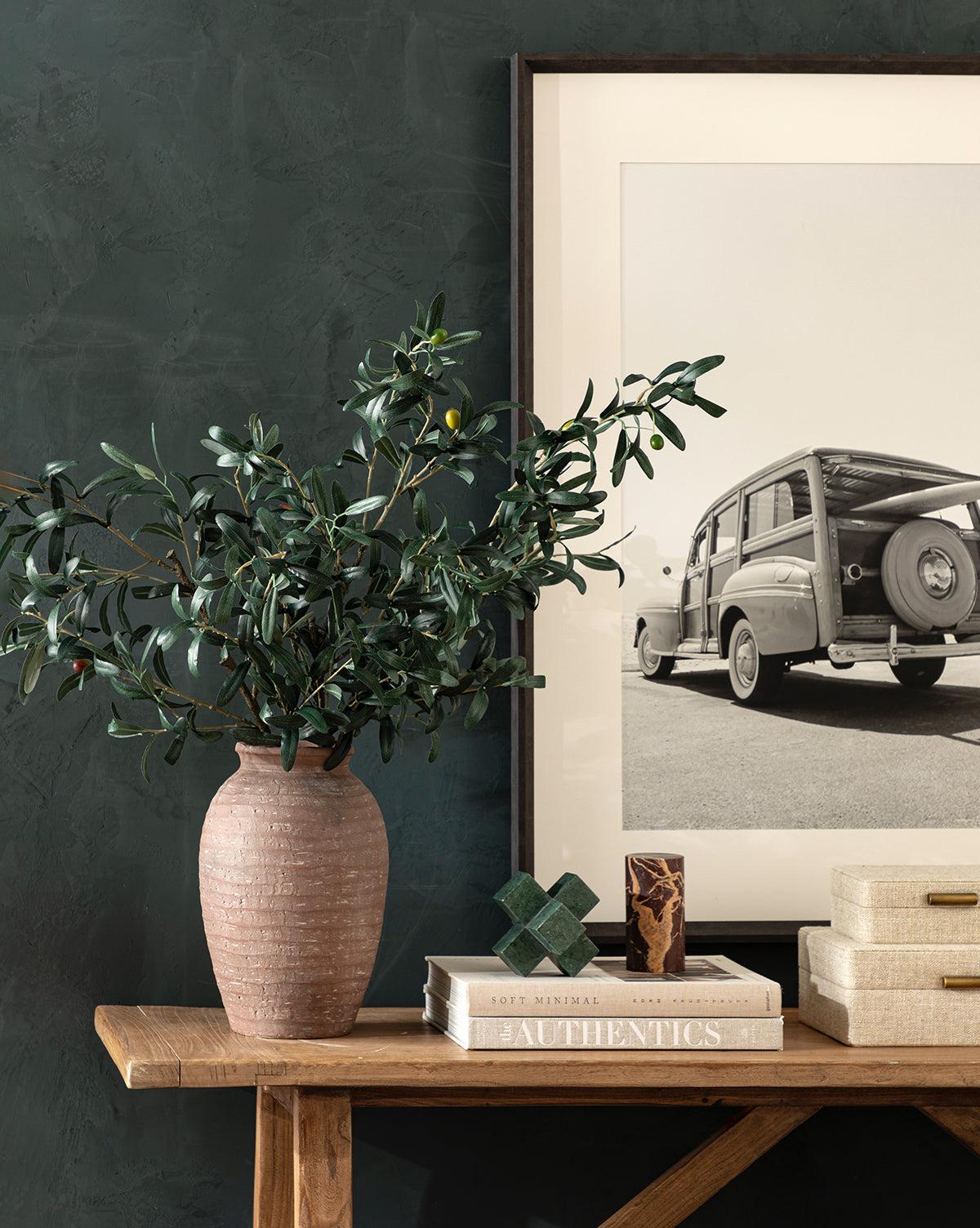 A wooden table with the Queta Terracotta Vase holding leafy branches, stacked books, decorative objects, and a framed black-and-white photo of a vintage station wagon against a dark textured wall.