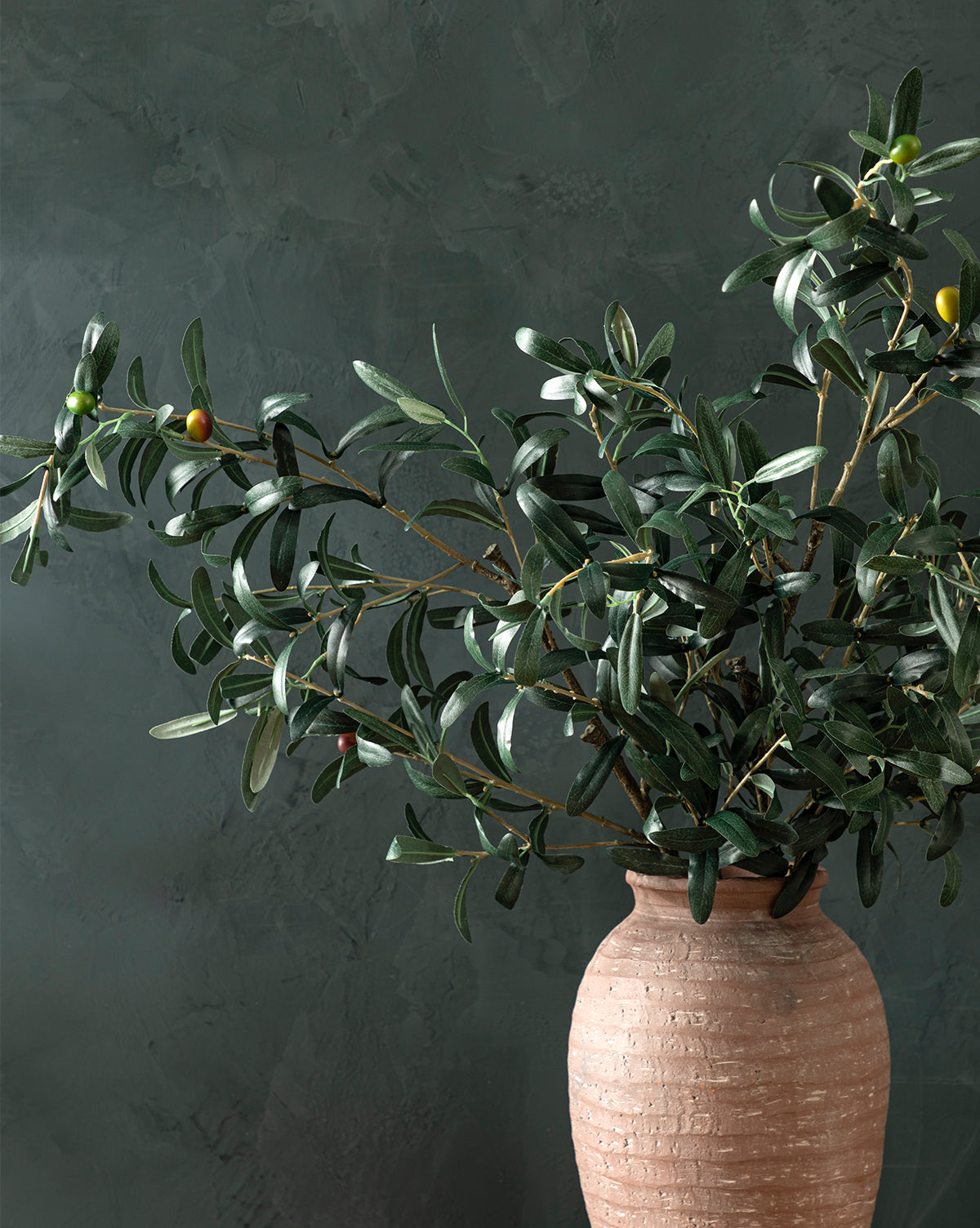 A Faux Olive Branch with dark green leaves and ripening olives is arranged in a terracotta vase, set against a textured dark green background, by McGee & Co.