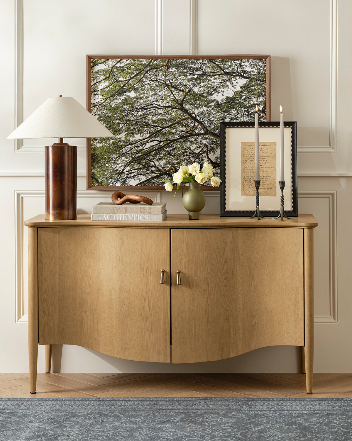 The Treetops giclee print, featuring tree branches, is displayed atop a wooden sideboard with curved doors. Also on the sideboard are a lamp, stacked books, white flowers in a vase, two tall candles, and a document.