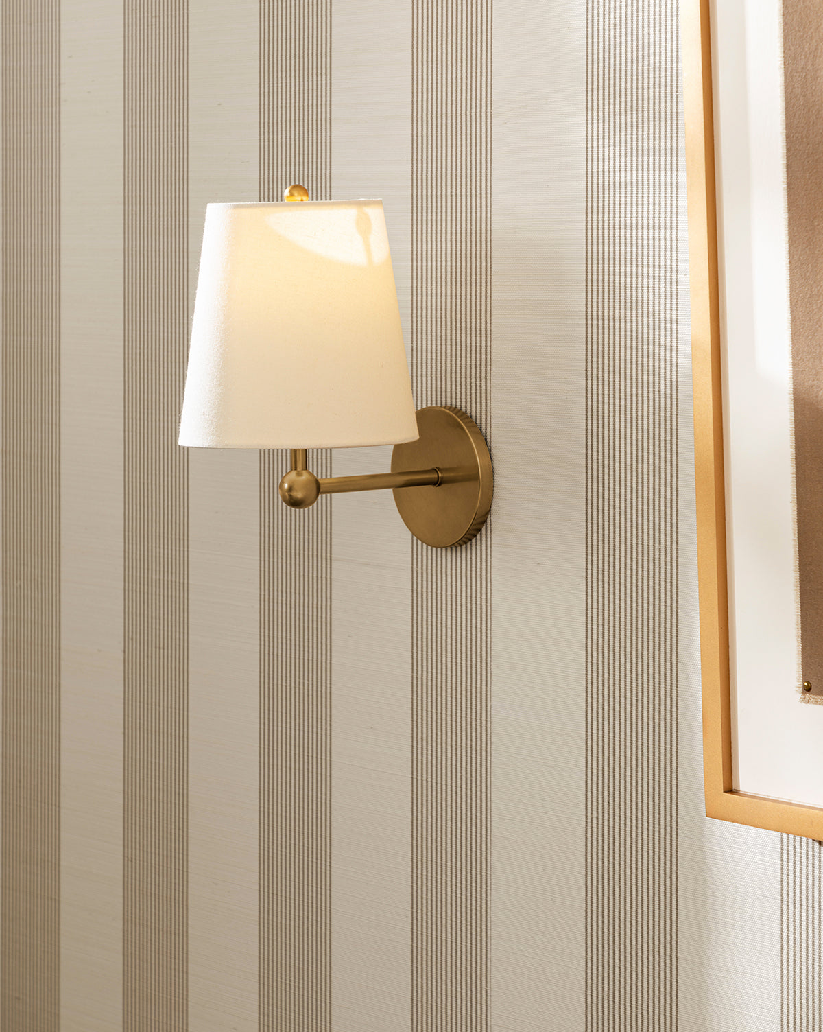 A brass wall-mounted lamp with a white shade glows against Leah Striped Sisal Wallpaper by York Wallcoverings. A gold-framed picture is partially visible on the right side of the image, McGee & Co.