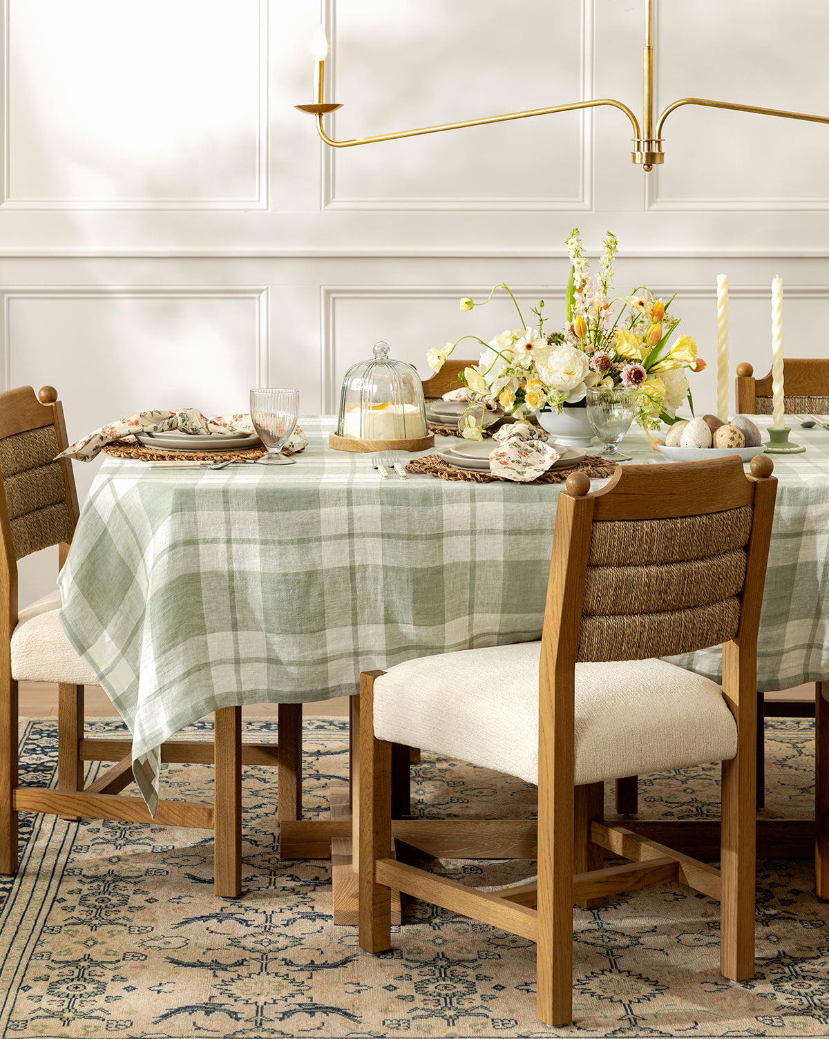 A dining table with a green and white checkered tablecloth is set for spring, surrounded by Alberta Dining Chairs on a patterned rug, with dishes, glasses, candles, flowers, and a cake under a glass dome, by McGee & Co.