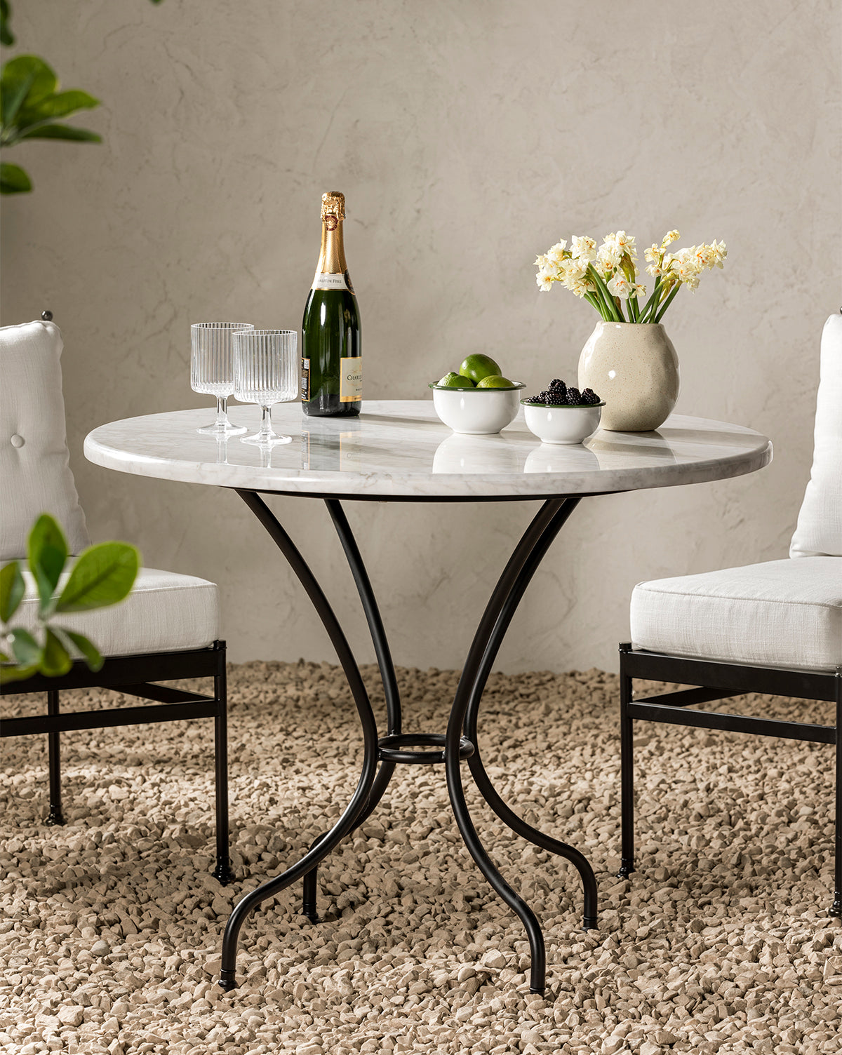 A round marble table with black curved legs stands on a gravel floor. On the table are two glasses, a bottle of champagne, a vase of daffodils, and Delia Bowls (Set of 3) filled with limes and blackberries. Two white cushioned chairs flank the table, by McGee & Co.
