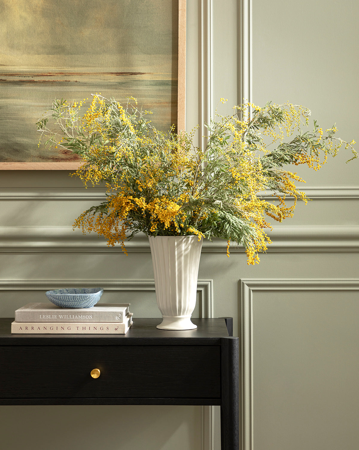 A Delphi Footed Vase filled with yellow and green wildflowers sits on a dark wood table beside a small bowl and stacked books, in front of a sage green paneled wall and framed painting, by McGee & Co.