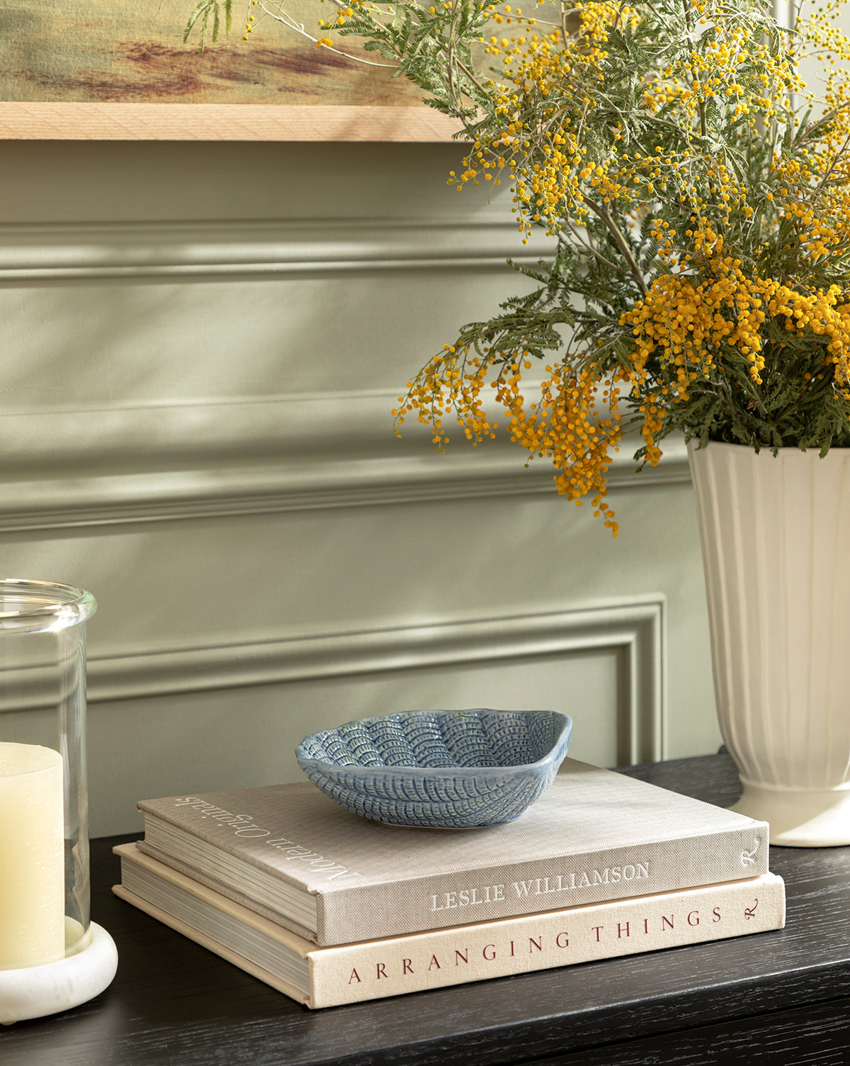 A decorative table features an Aurelie Shell Shaped Bowl in coastal blue stoneware atop two stacked books, accompanied by a glass candle and a white vase with yellow flowers, all set against a sage green paneled wall, by McGee & Co.