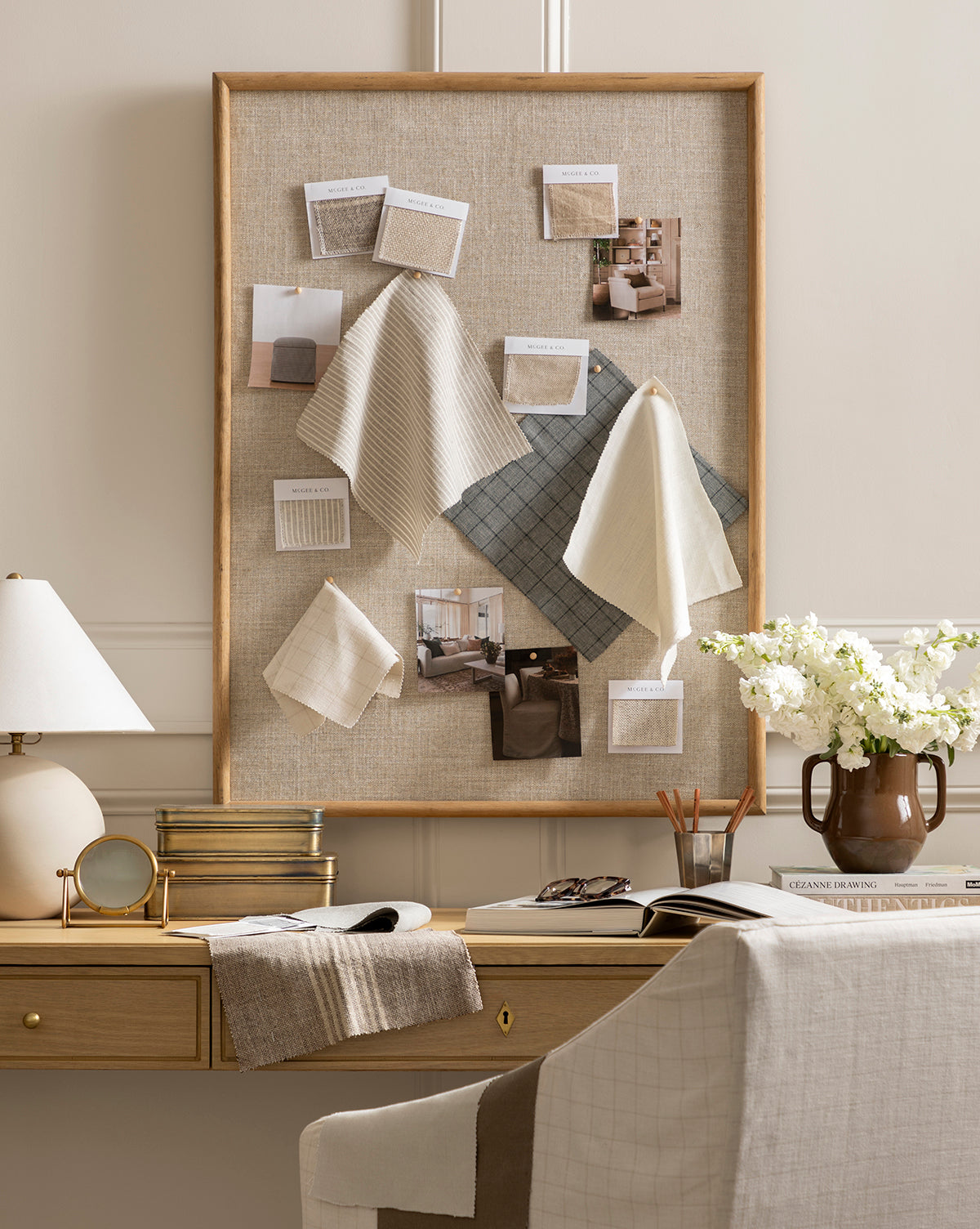 The Oak & Linen Memo Board showcases fabric swatches and design photos above a wooden desk with a lamp, vases, flowers, and notebooks. A fabric-draped chair completes this stylish and organized office space.
