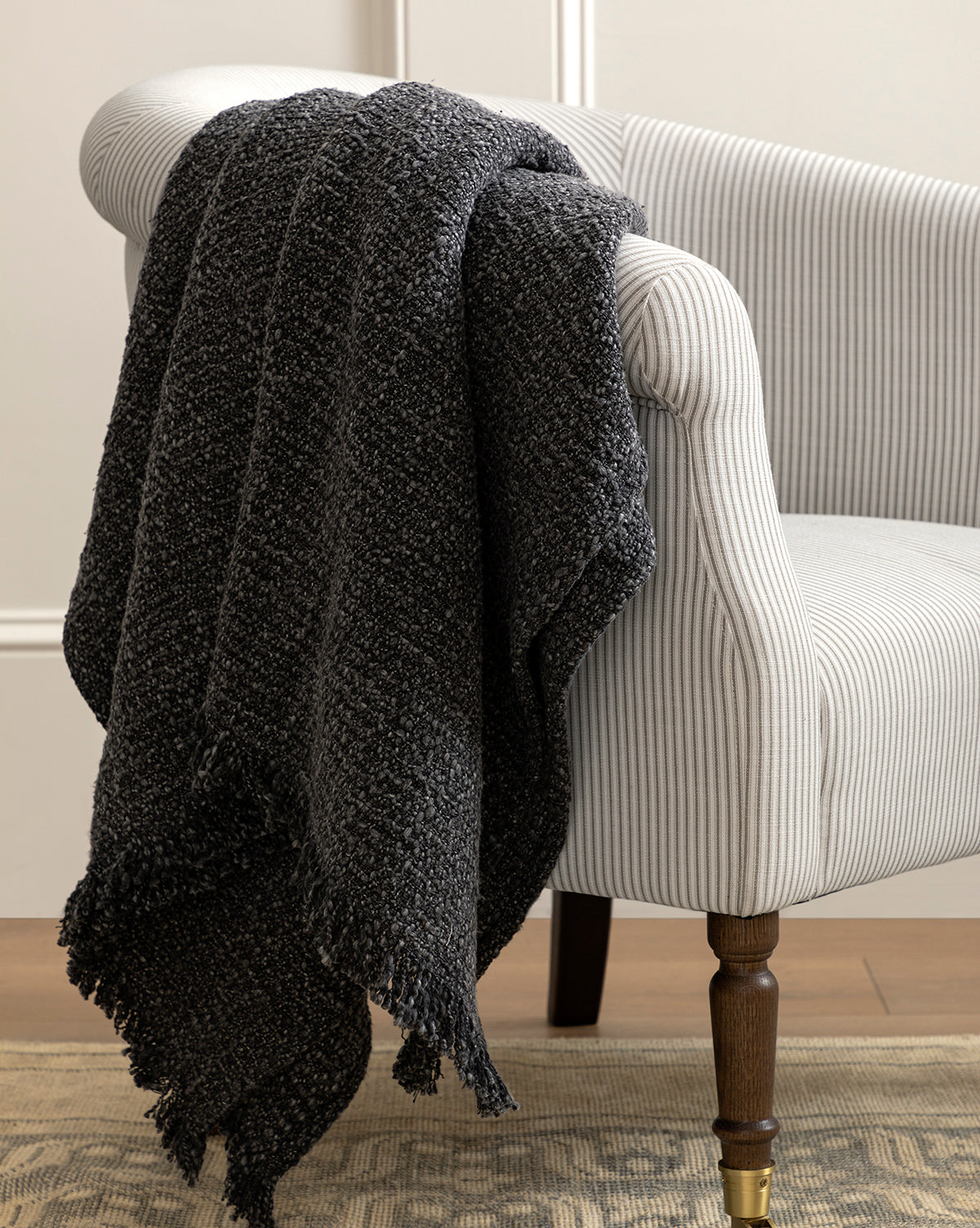 The Clarissa Throw Blanket by Creative Co-op, featuring a soft boucle texture and charcoal hue, is draped over the arm of a white and gray pinstriped chair with wooden legs, all set atop a patterned area rug. McGee & Co.