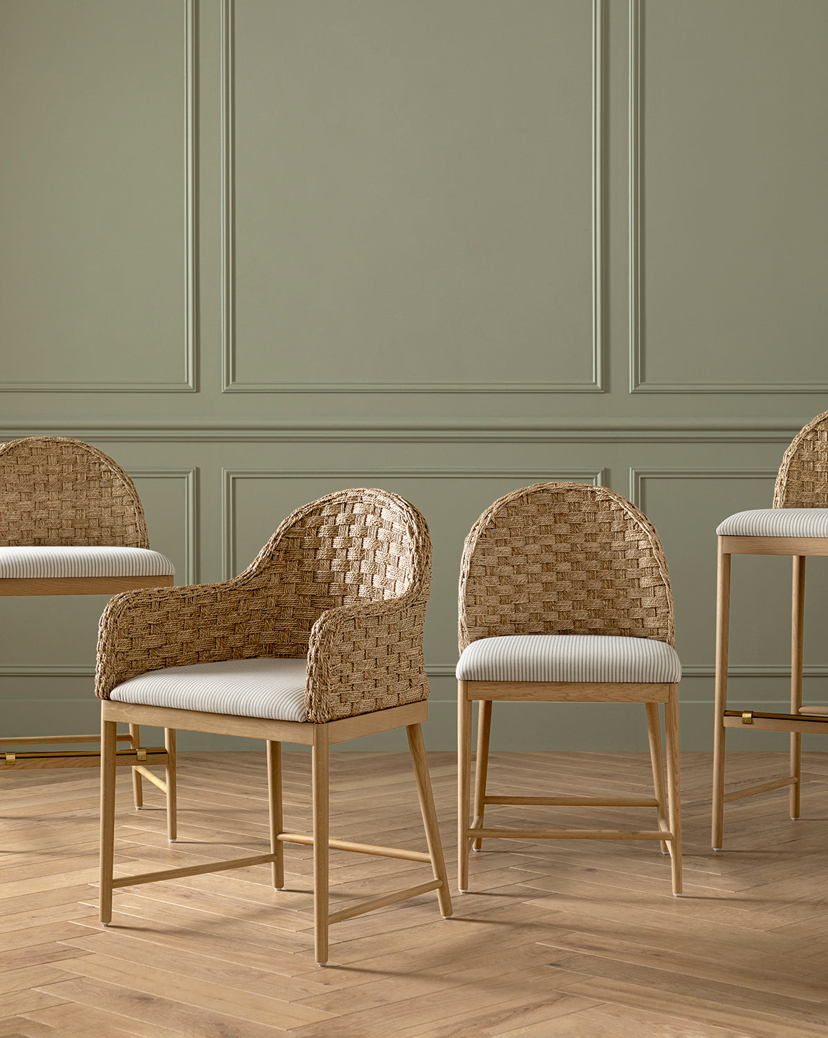 Four Molly Dining Chairs with woven rattan seats and solid oak legs are set on a wooden floor before a sage green paneled wall; two feature armrests and two are taller bar stools, by McGee & Co.
