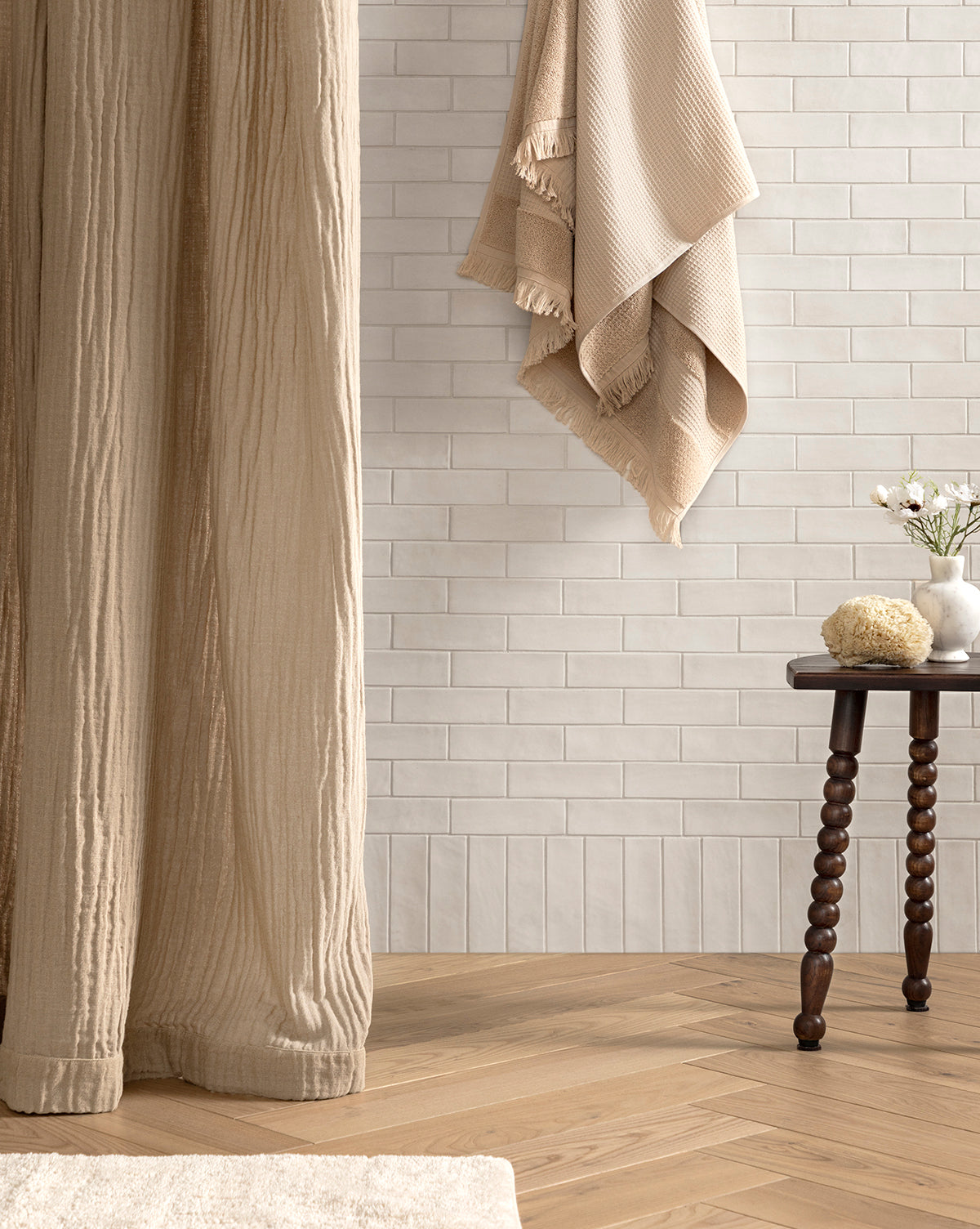The Mirella Shower Curtain in beige hangs beside a small wooden table topped with a sponge, vase of flowers, and glass. White subway tiles serve as the backdrop, complemented by light brown herringbone flooring.