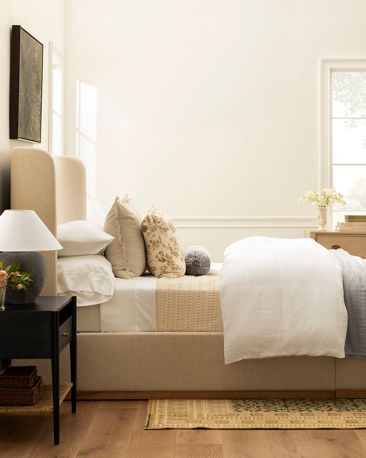 A cozy, sunlit bedroom features a beige upholstered bed with white and patterned pillows, 100% cotton bedding, and the Carnel Waffle Blanket from Portugal for texture. A black nightstand with flowers and books sits beside two large windows.