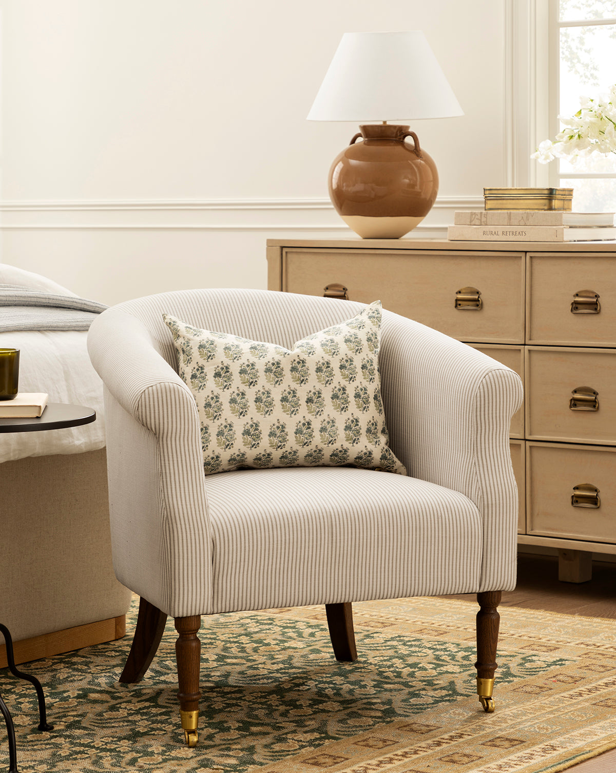 The Makerspalm Clemence Lounge Chair with a patterned pillow sits on a patterned rug beside a wooden dresser topped with a ceramic lamp, books, and flowers in a cozy, well-lit bedroom, McGee & Co.