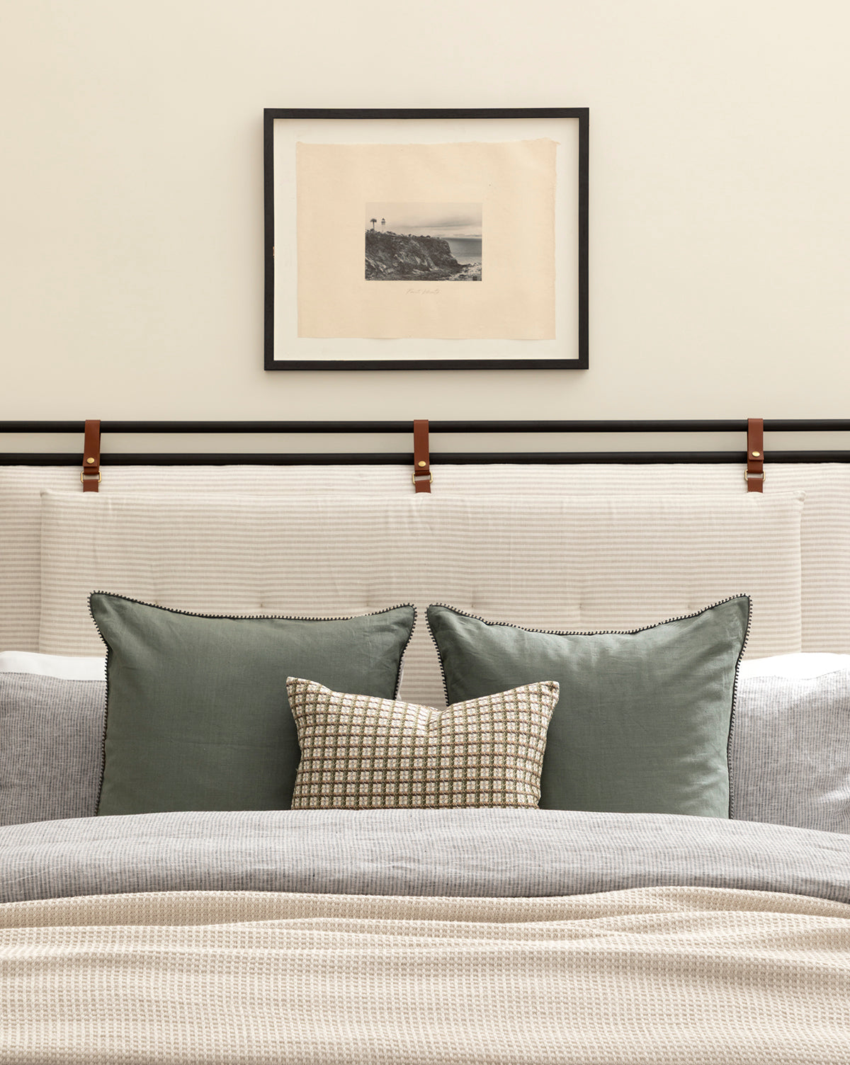 The Point Vicente bed features a light padded headboard with leather straps, green and patterned pillows, a textured blanket, and sits beneath framed vintage wall art on a beige wall.