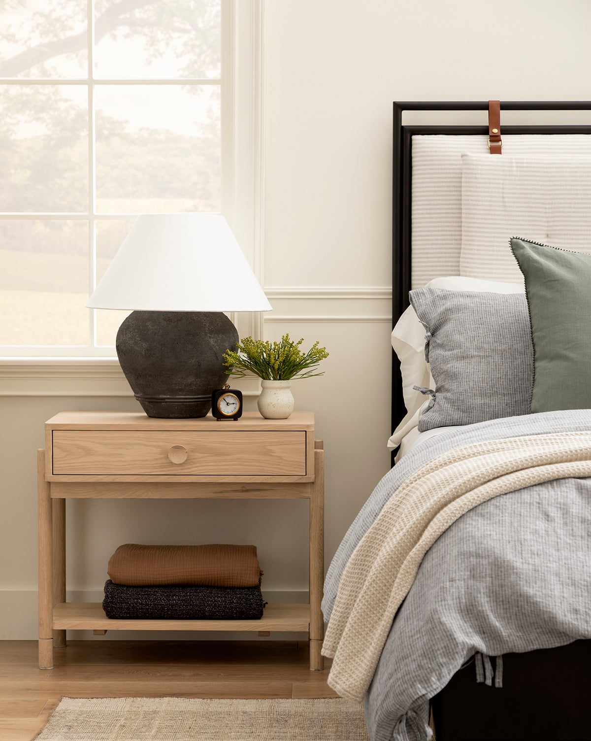 A wooden nightstand with a black lamp, small plant, Kya Clock, and ceramic bowl sits beside a bed made with gray and white bedding, a green pillow, and a window in the background, by McGee & Co.