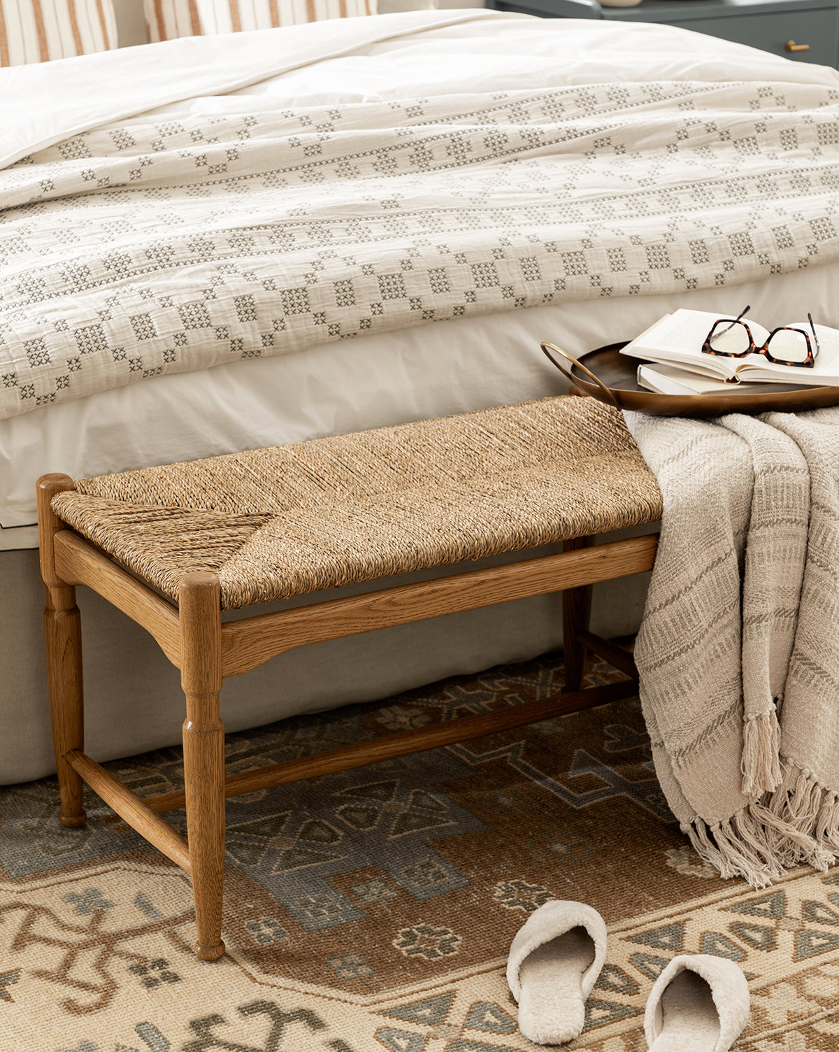 The Phipps Woven Bench with a seagrass seat and beige throw blanket sits at the foot of a bed with patterned bedding. A tray with a book and glasses rests on the bench, while slippers sit nearby on a patterned rug, by McGee & Co.