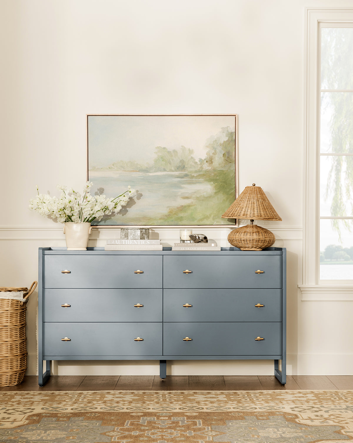 A blue dresser with gold handles stands against a white wall, topped with a lamp, books, and white flowers. Above it hangs Lake by Lauren Fuhr in a maple floater frame; nearby are a window and woven basket.