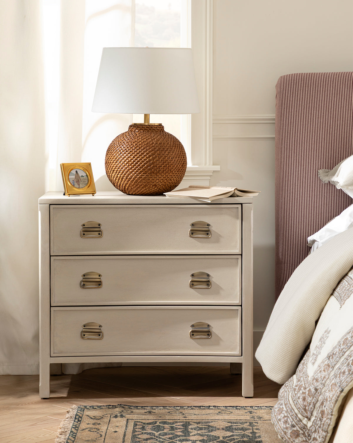 A beige nightstand with three drawers holds the Avedon Table Lamp with a natural rattan base and white linen shade, a small framed photo, and an open book. Beside it sits a bed dressed in neutral bedding and topped with a red-striped upholstered headboard by McGee & Co.