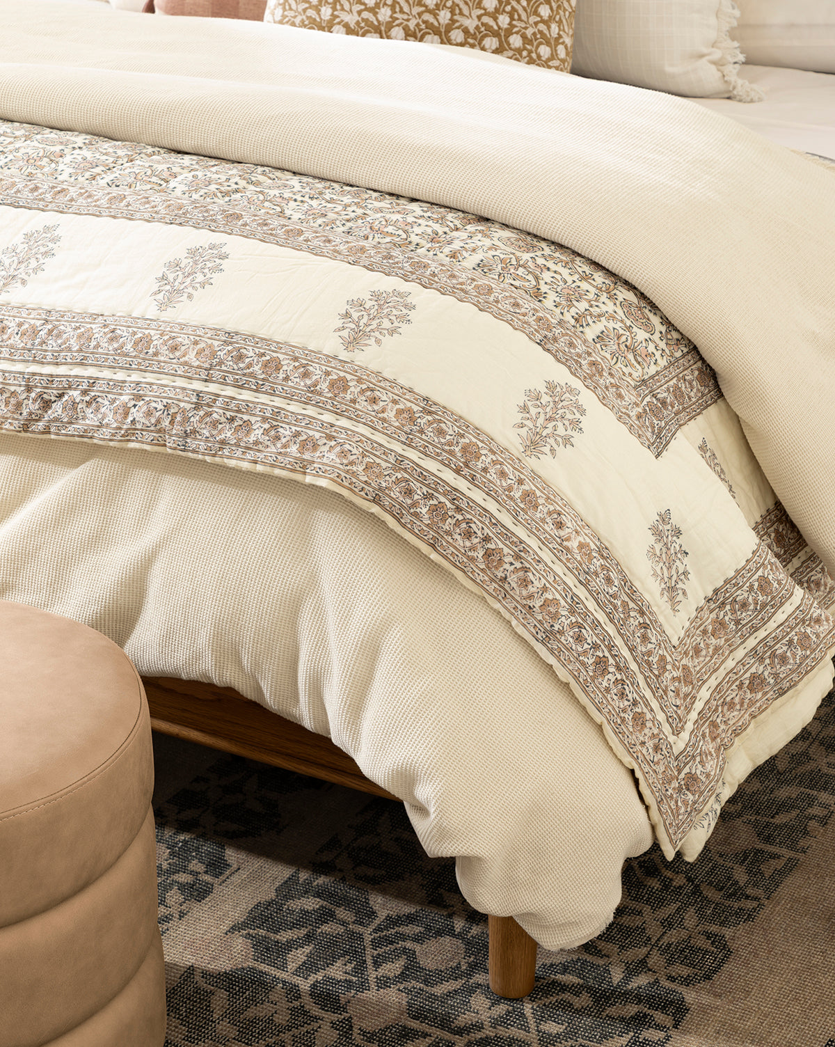 A close-up of a neatly made bed features the Leander Quilt in cream and beige cotton, paired with a textured beige comforter and decorative pillows on a wooden frame, all atop a floral-patterned rug for inviting charm.