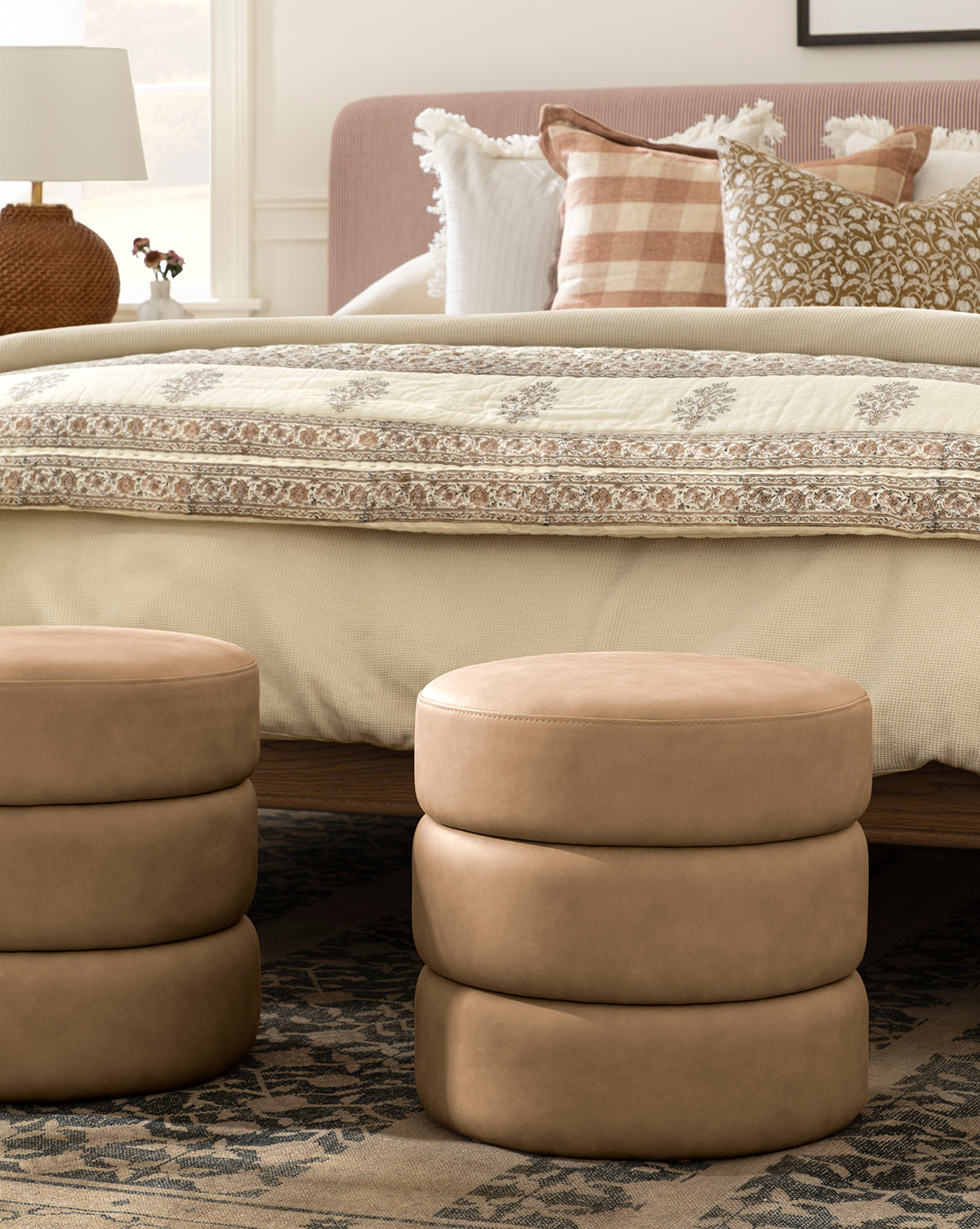 A cozy bedroom features a bed with patterned pillows and a quilted blanket, flanked by two round, tan Channel Ottomans on a patterned rug. A lamp and small decor accent the nightstand beside the bed, by McGee & Co.