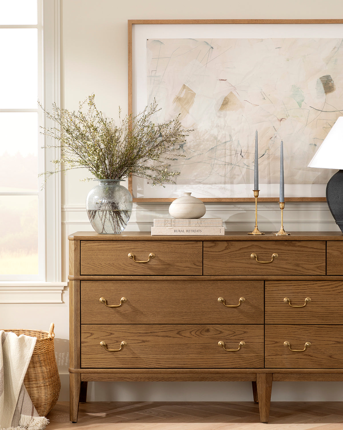 A wooden dresser with gold handles displays the Gretchen Vase filled with branches, a small round ceramic object, two tall candlesticks, and a lamp. Abstract art hangs above as sunlight pours through a nearby window, by McGee & Co.