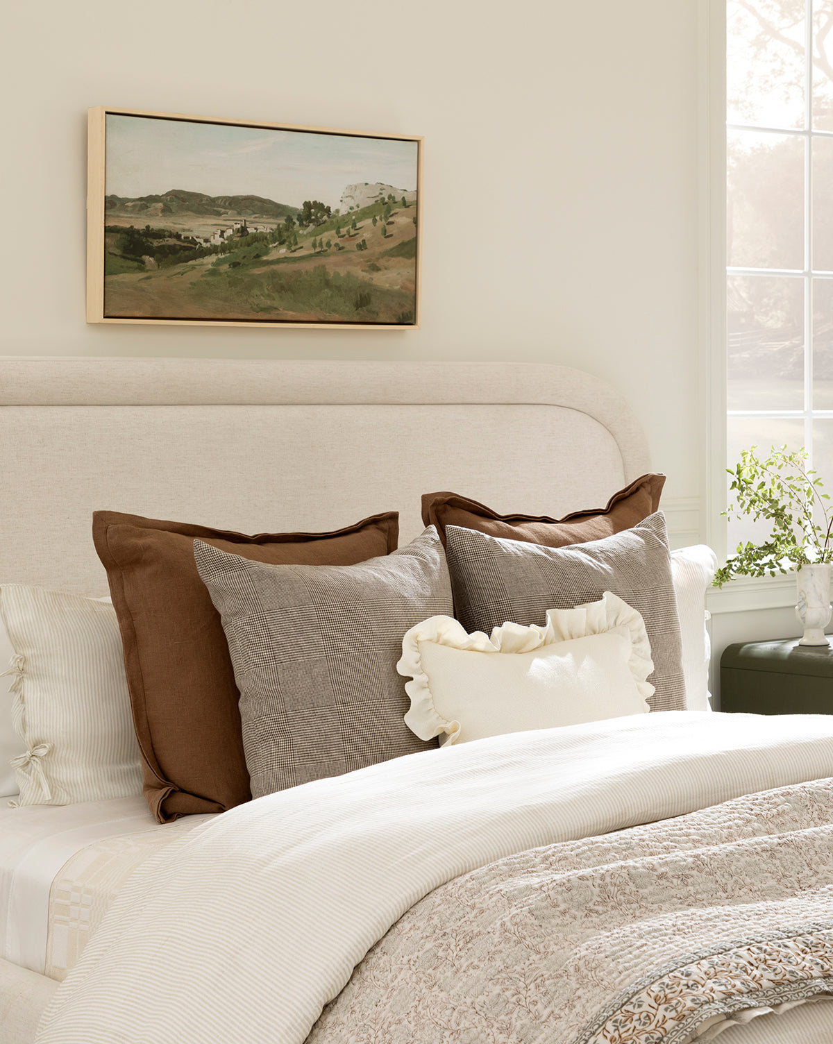 A neatly made bed with neutral bedding, large brown and gray pillows, and a ruffled decorative pillow sits below a padded headboard. Above it hangs the View of the Town canvas. Sunlight streams in past a window with a small plant on the sill, by McGee & Co.