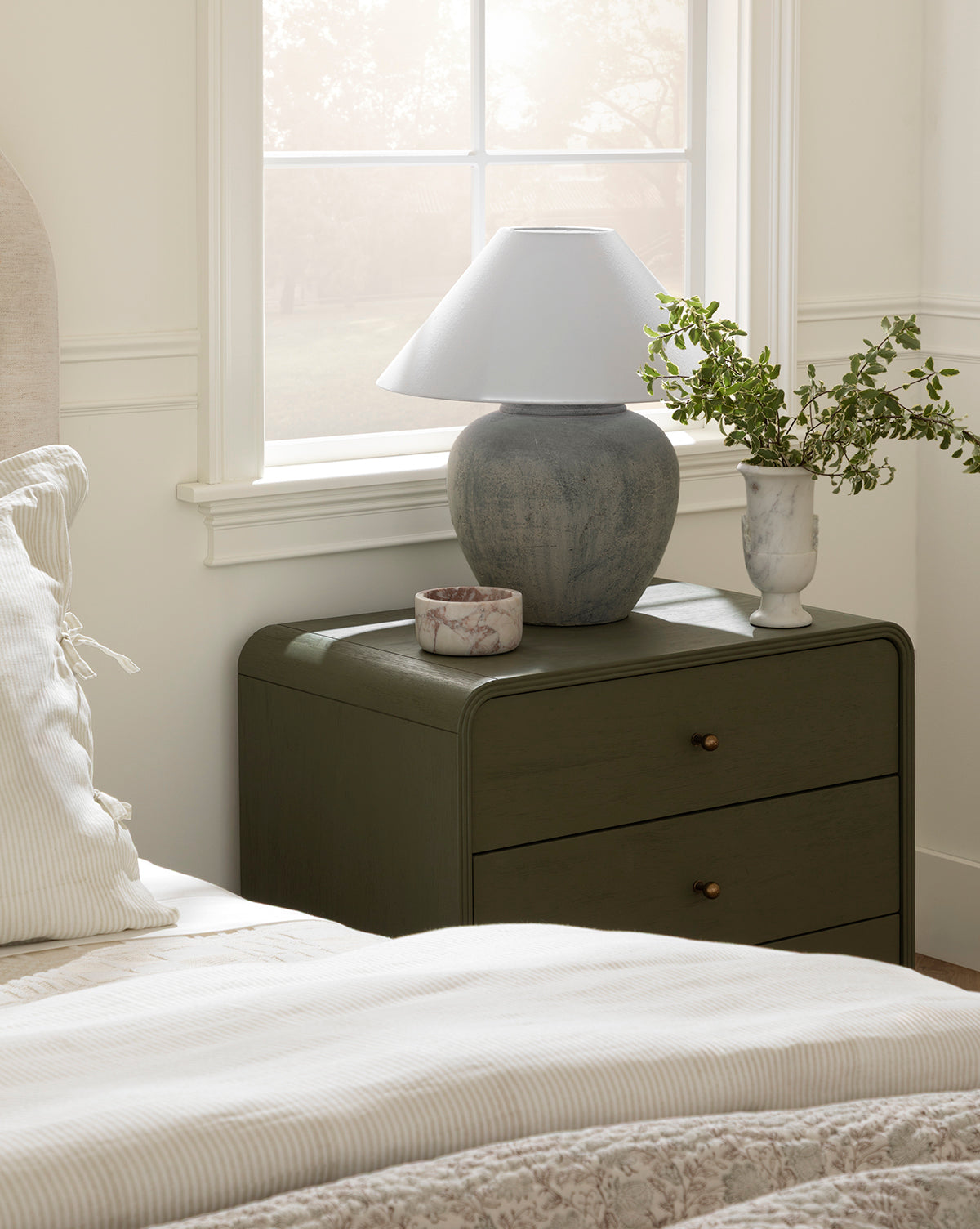 A bedside table holds a lamp, a small purple and white dish, and the Rayla Marble Vase with greenery next to a bed with light bedding. Sunlight streams through the window, adding calm, cozy vibes inspired by traditional decor, by McGee & Co.