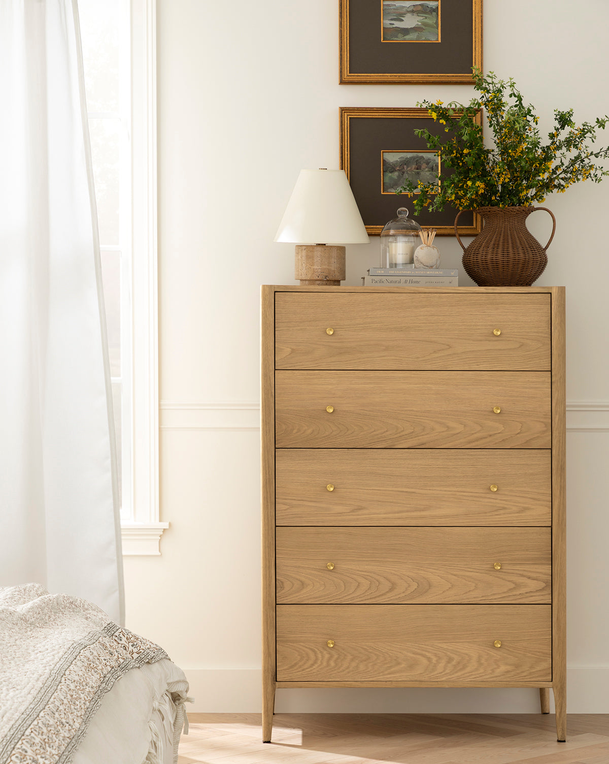 A light wood dresser with four drawers and gold knobs stands by a white wall. On top are a brown vase with green branches, the Bronte Table Lamp, and framed art above. Sunlight streams in from a nearby window by McGee & Co.