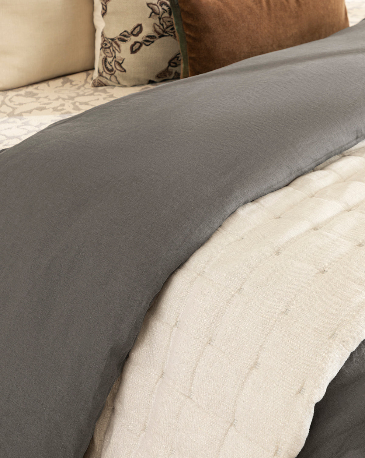 A close-up of a neatly made bed with the Chaucer Quilt, featuring gray quilted stitching and off-white linen-cotton texture, accented by decorative cream and brown floral-embroidered pillows.